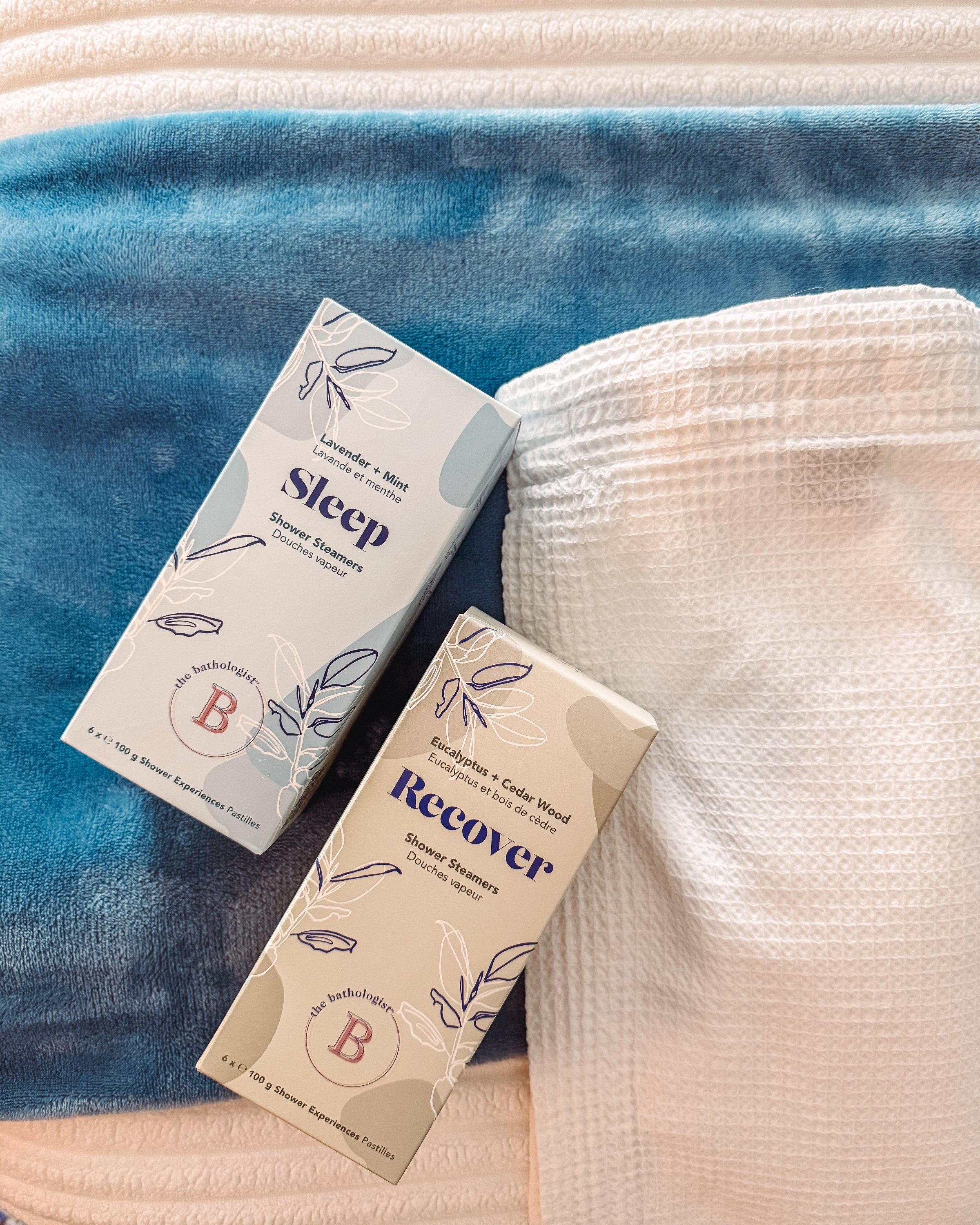Two product boxes labeled 'Sleep' and 'Recover' placed on a soft blue blanket next to a white towel.