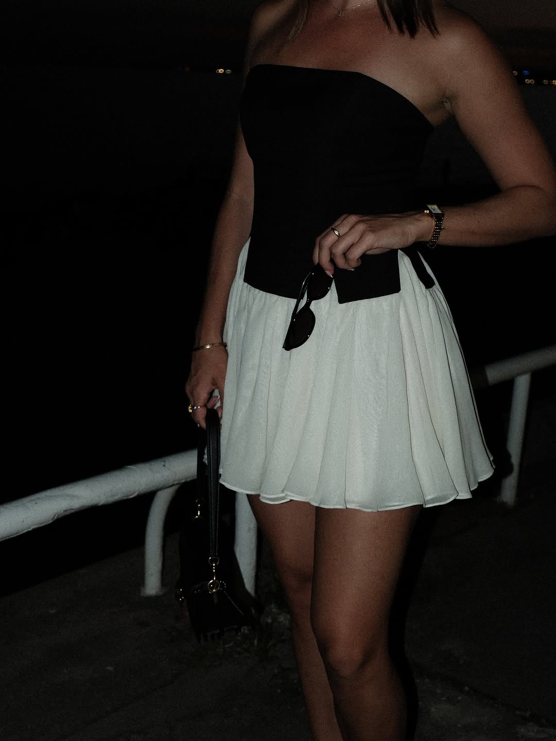 A person wearing a strapless black top and a white skirt stands near a waterfront at dusk, holding sunglasses and a small handbag.