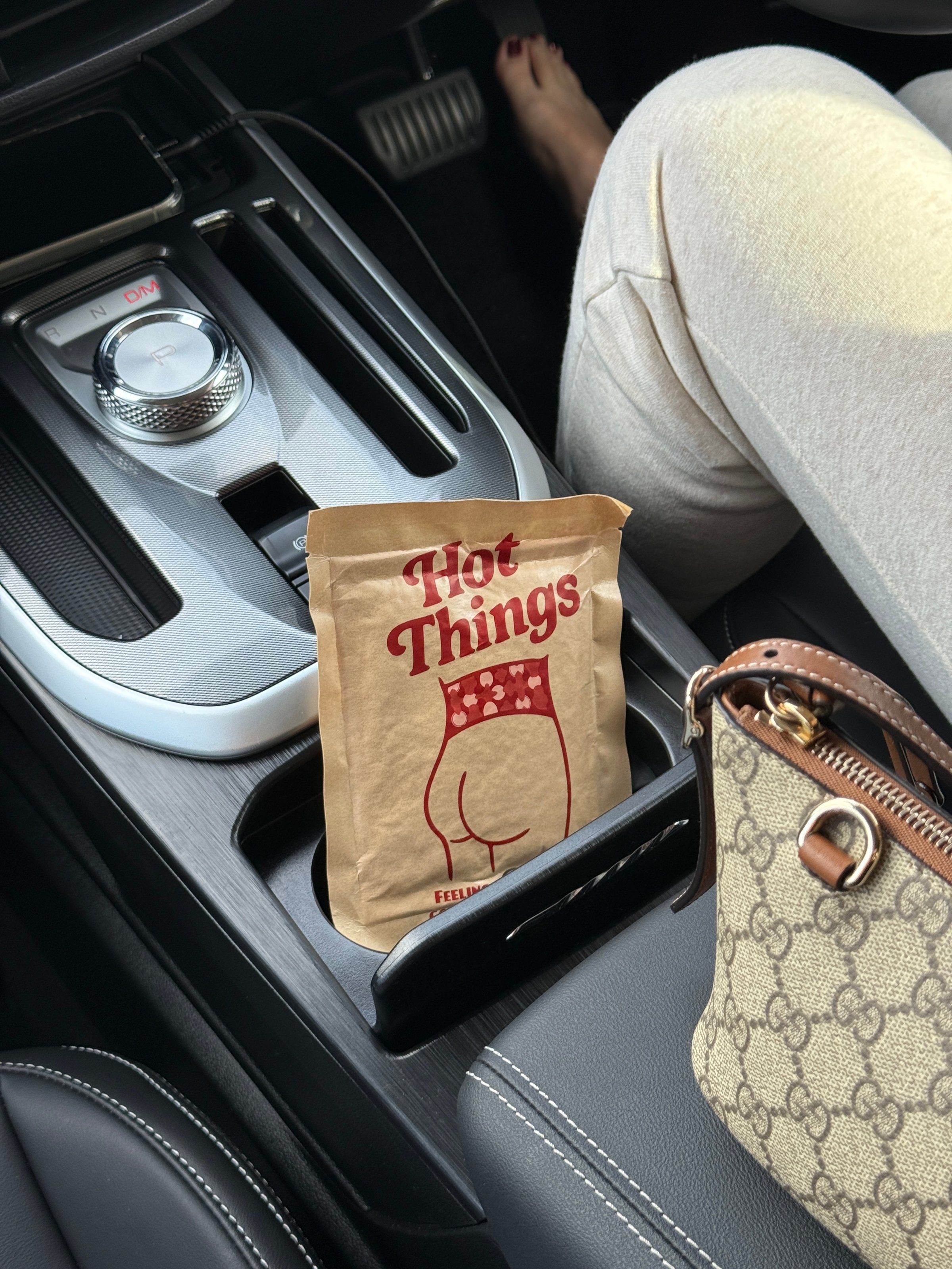 A humorous snack bag labeled 'Tiot Things' placed in the center console of a car, with a person's leg visible in light-colored pants.