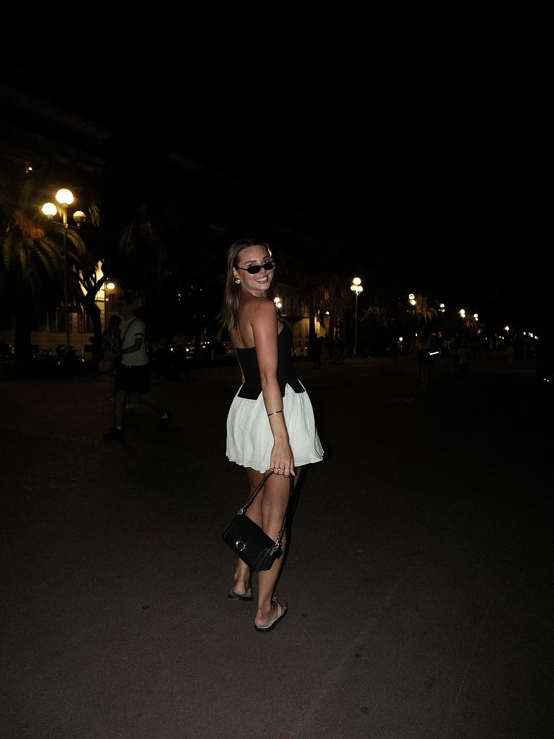 A woman poses in a stylish outfit on a nighttime street, illuminated by streetlights and palm trees in the background.