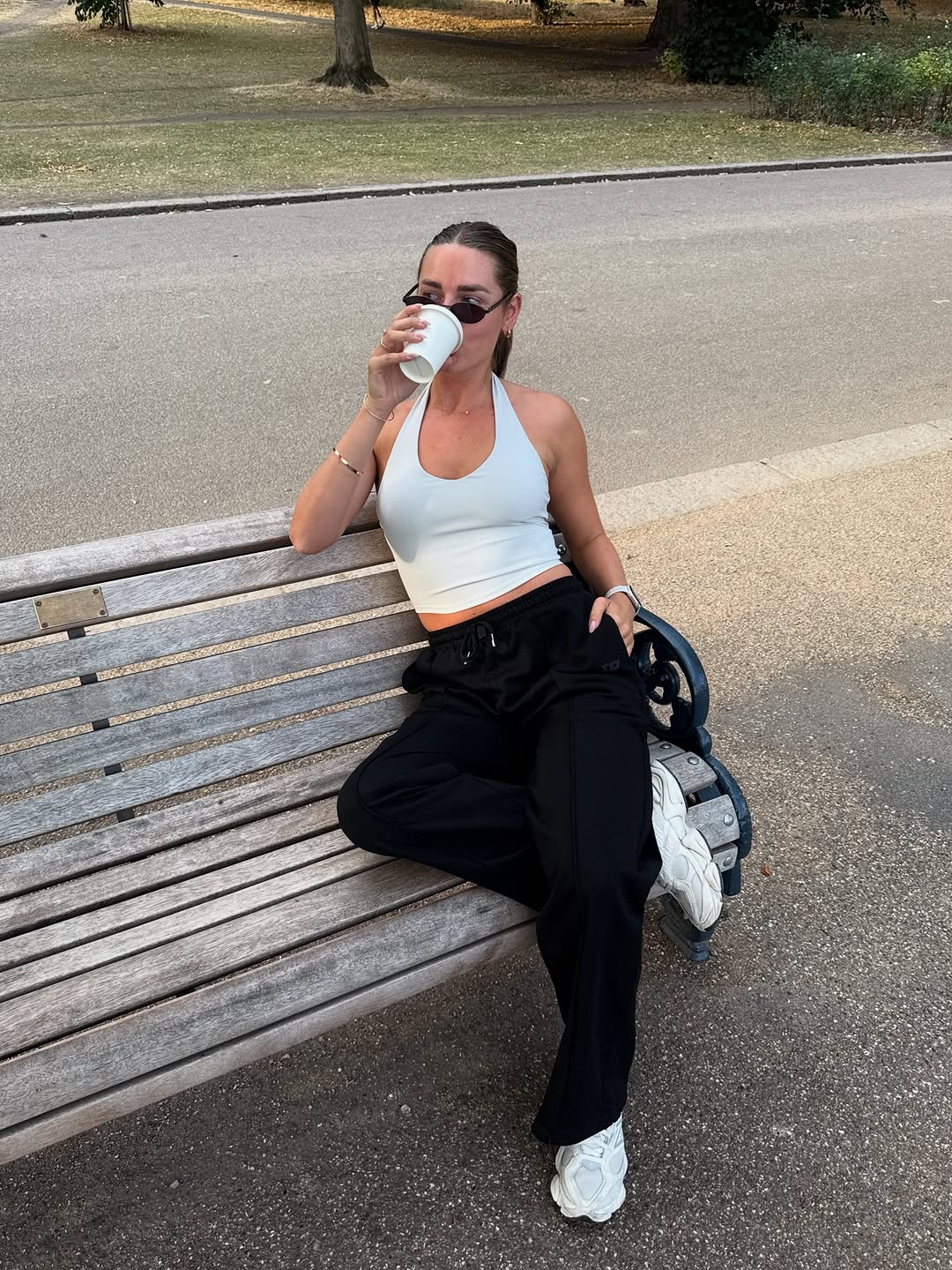 A person sitting on a bench in a park, wearing a white crop top and black pants, sipping from a coffee cup.
