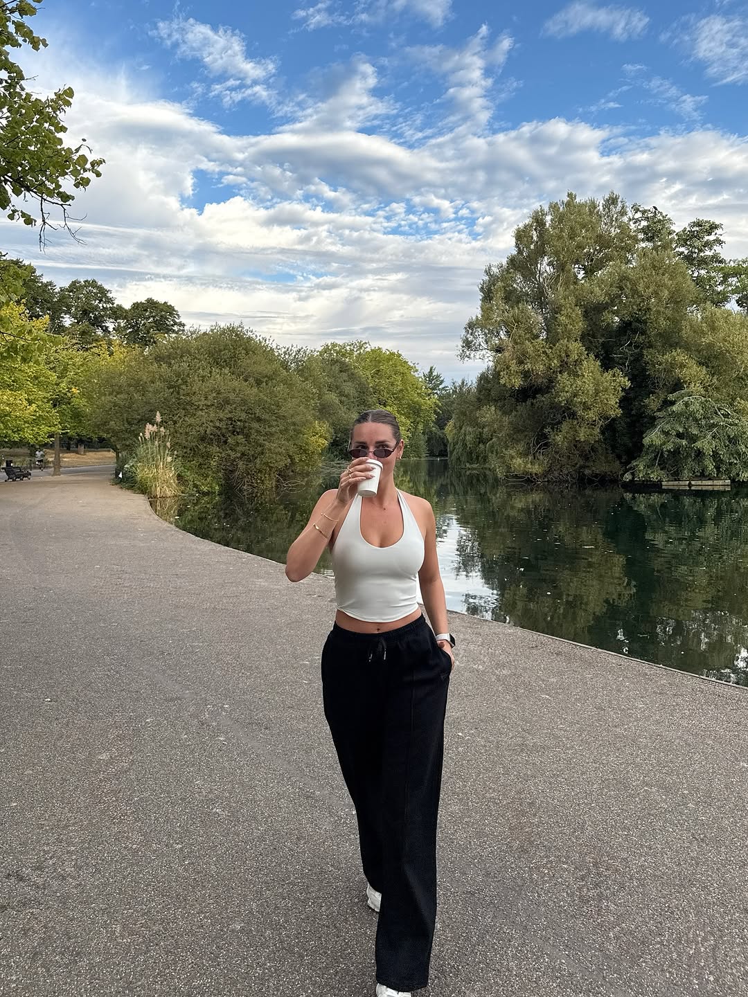 A woman in a white tank top and black pants stands by a serene body of water, sipping from a cup while surrounded by lush greenery and a blue sky.