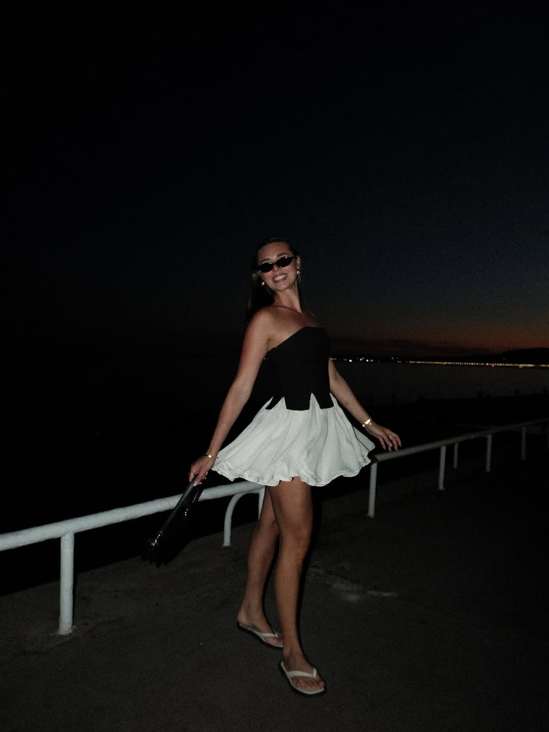 A woman in a stylish black and white dress poses on a walkway at dusk, with a scenic view of the horizon in the background.