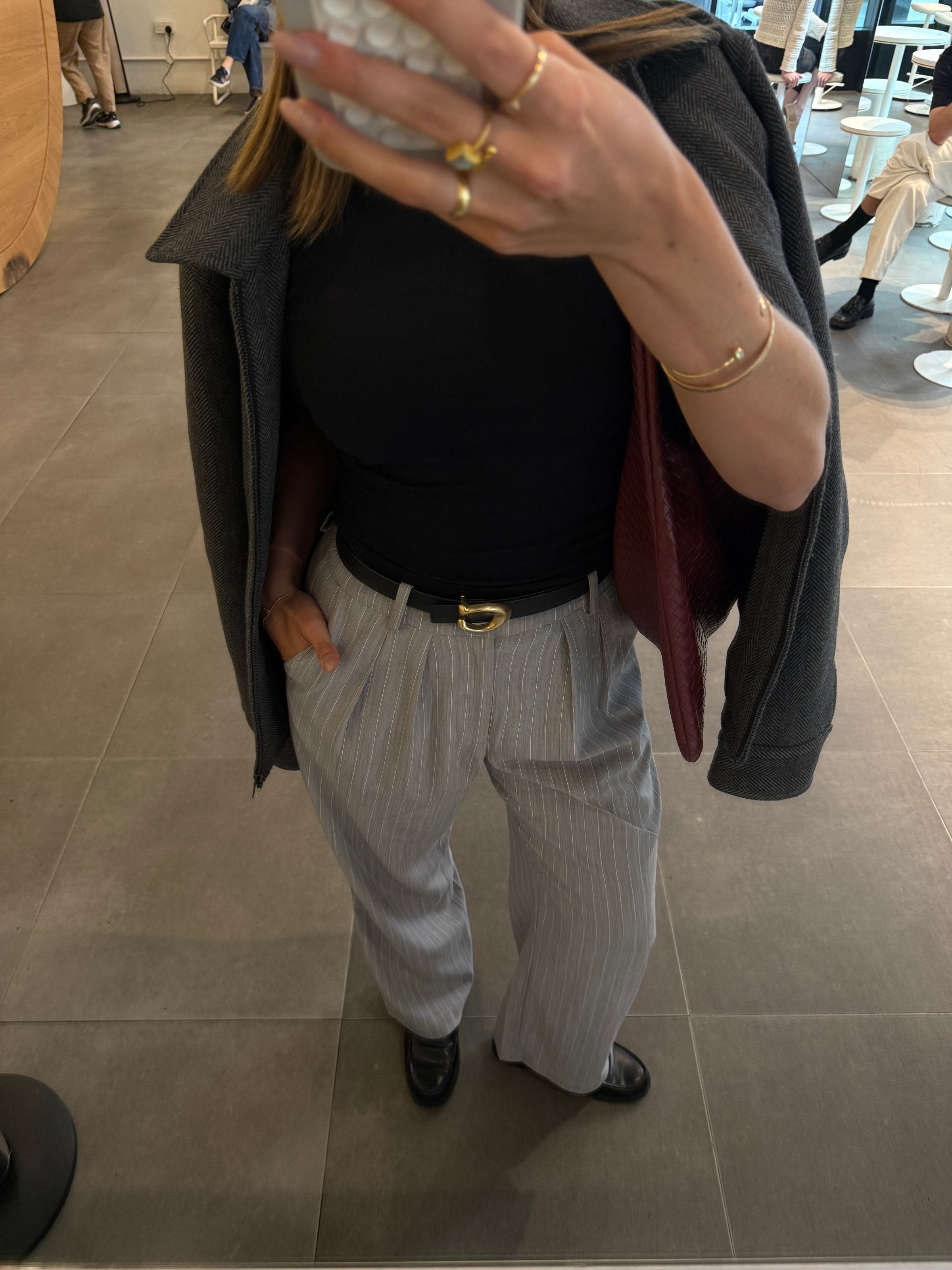 A person is taking a mirror selfie, wearing a black top and gray pinstripe pants, accessorized with rings and a belt.
