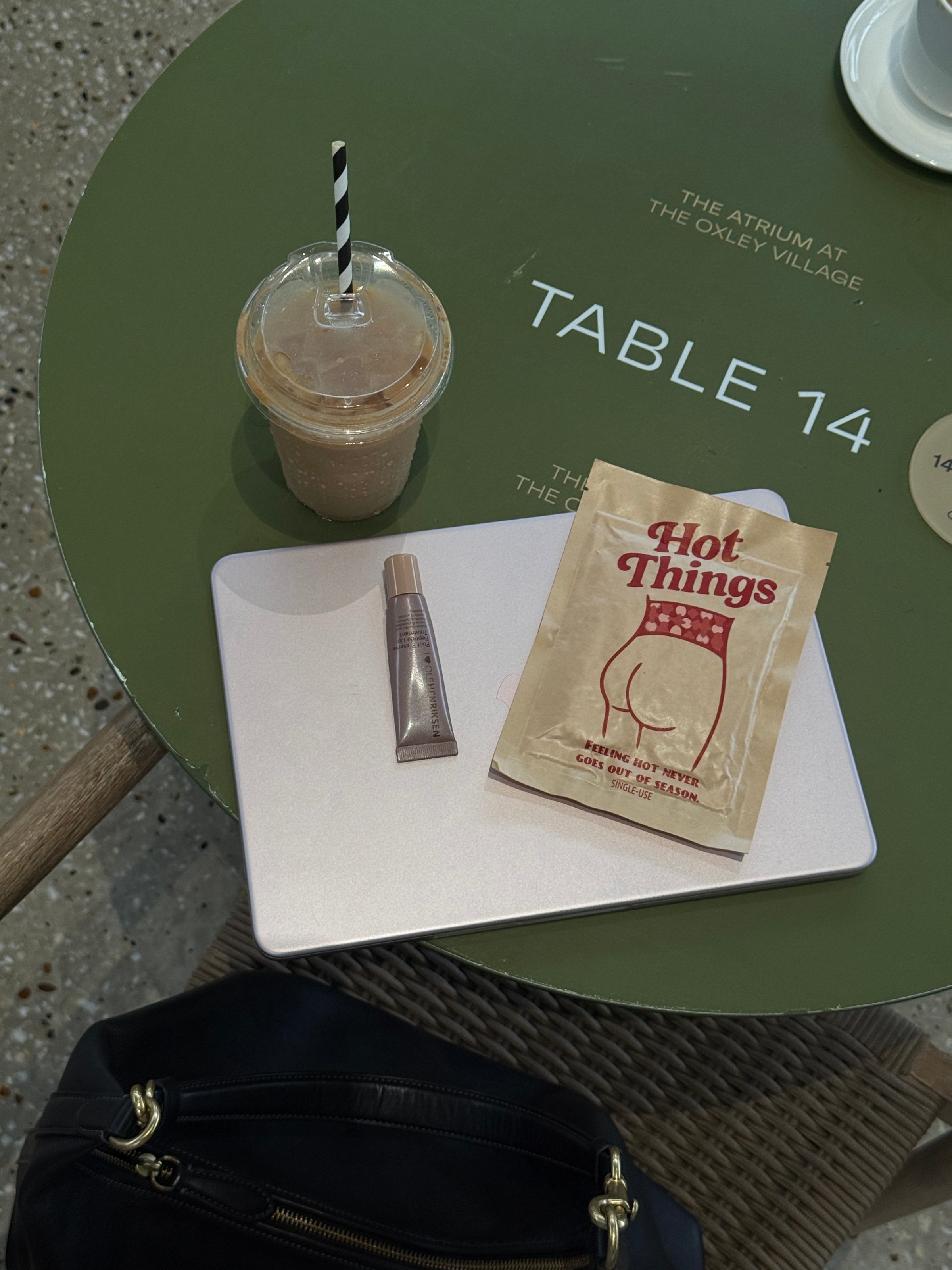A laptop and a makeup product are placed on a round green table, alongside a cup of iced coffee and a bag of snacks.
