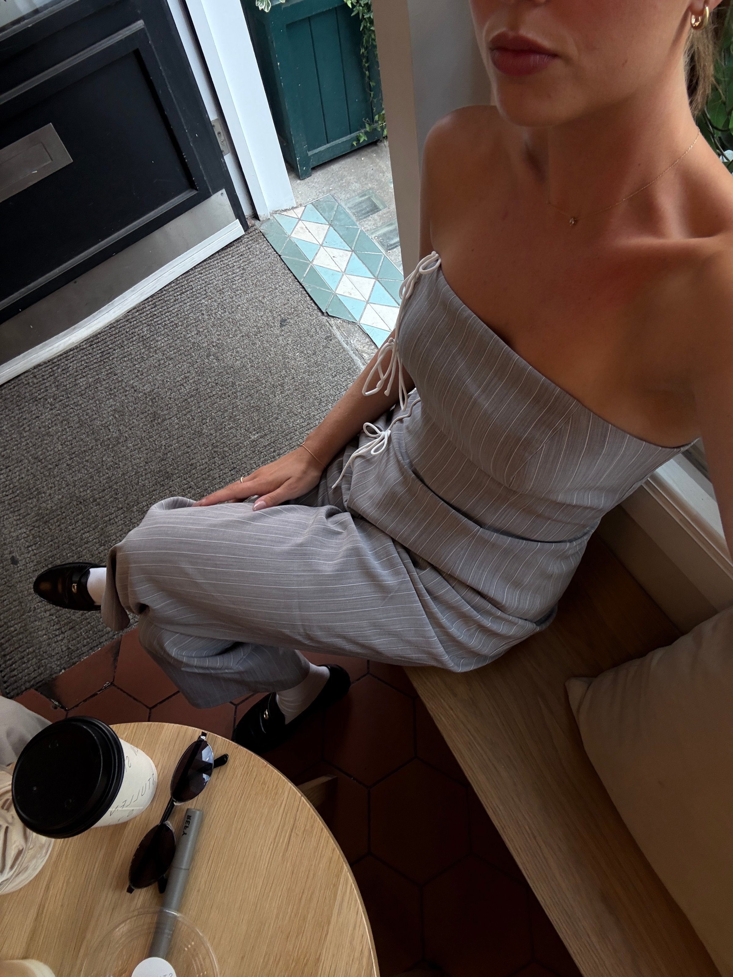 A woman sitting at a cafΓ© wearing a strapless striped jumpsuit, with a coffee cup and sunglasses on the table.