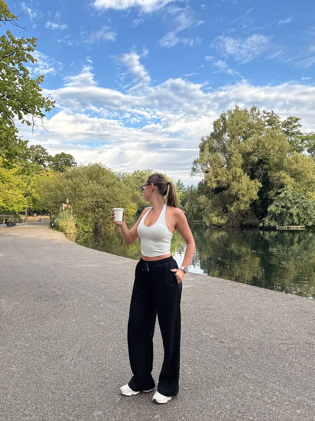 A person stands by a serene body of water, wearing a white tank top and black pants, holding a coffee cup while looking towards the sky.