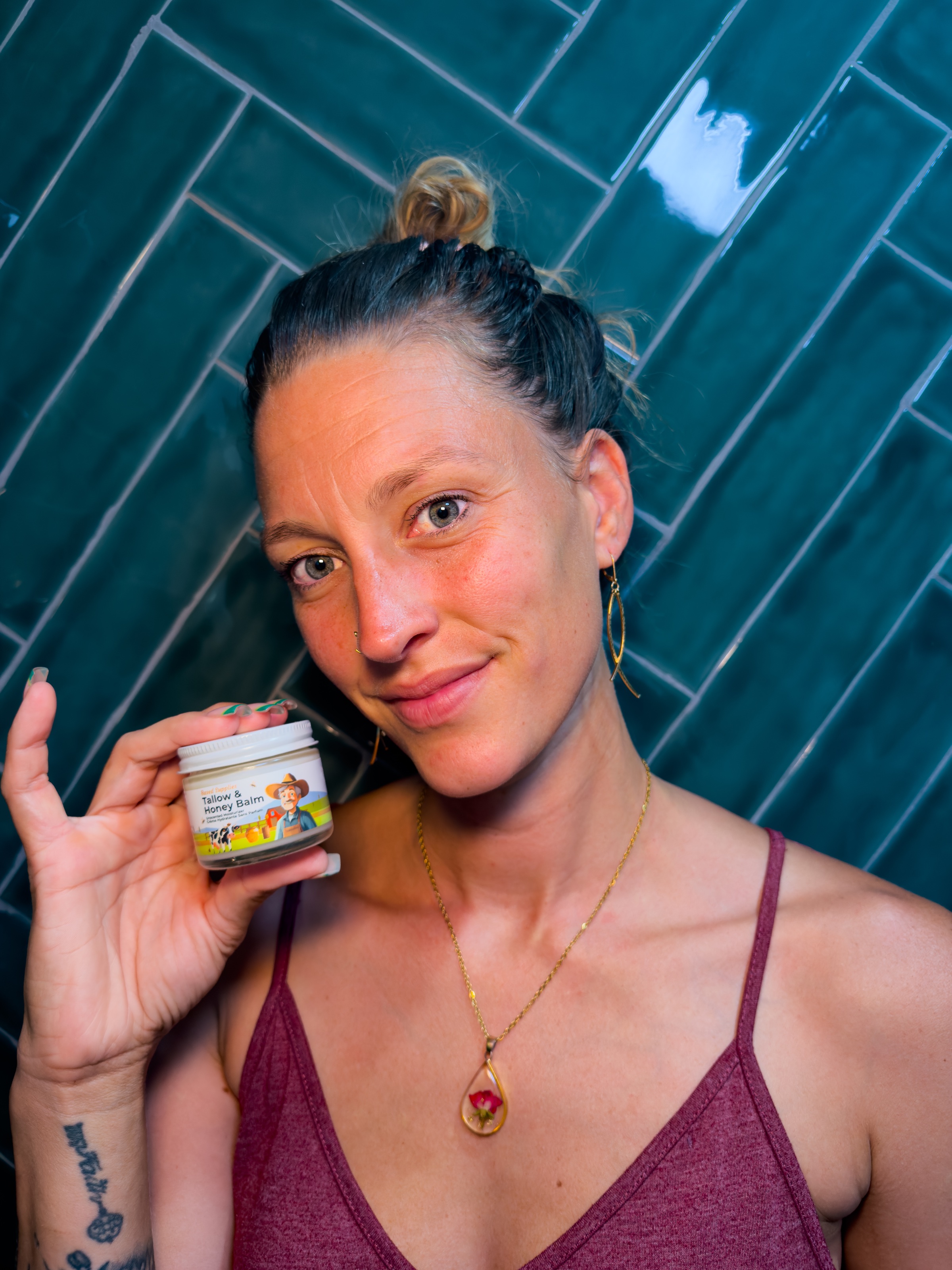 A woman holds a small jar of skincare product while smiling at the camera, with a tiled wall in the background.