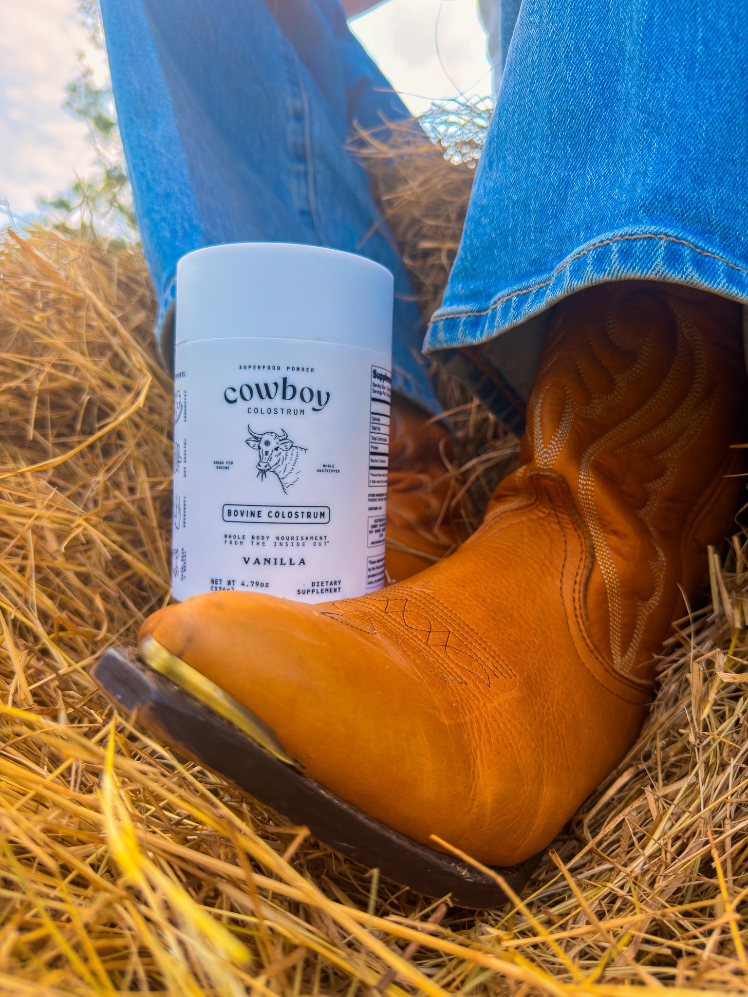 A close-up of a cowboy boot resting on hay, next to a container of cowboy-themed vanilla body lotion.