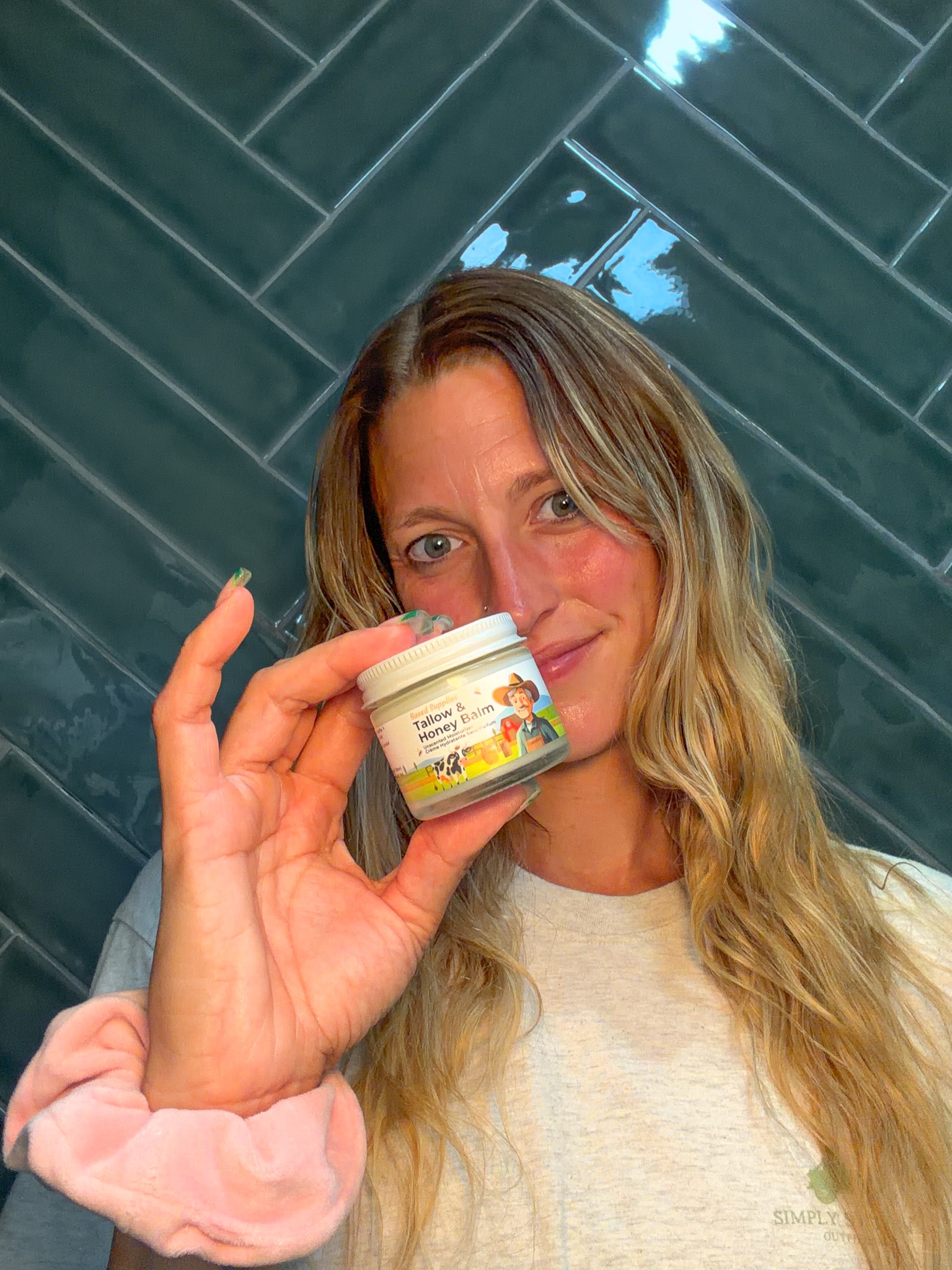 A woman with long, wavy hair holds a small jar of skincare product in front of her face, smiling. The background features glossy green tiles.
