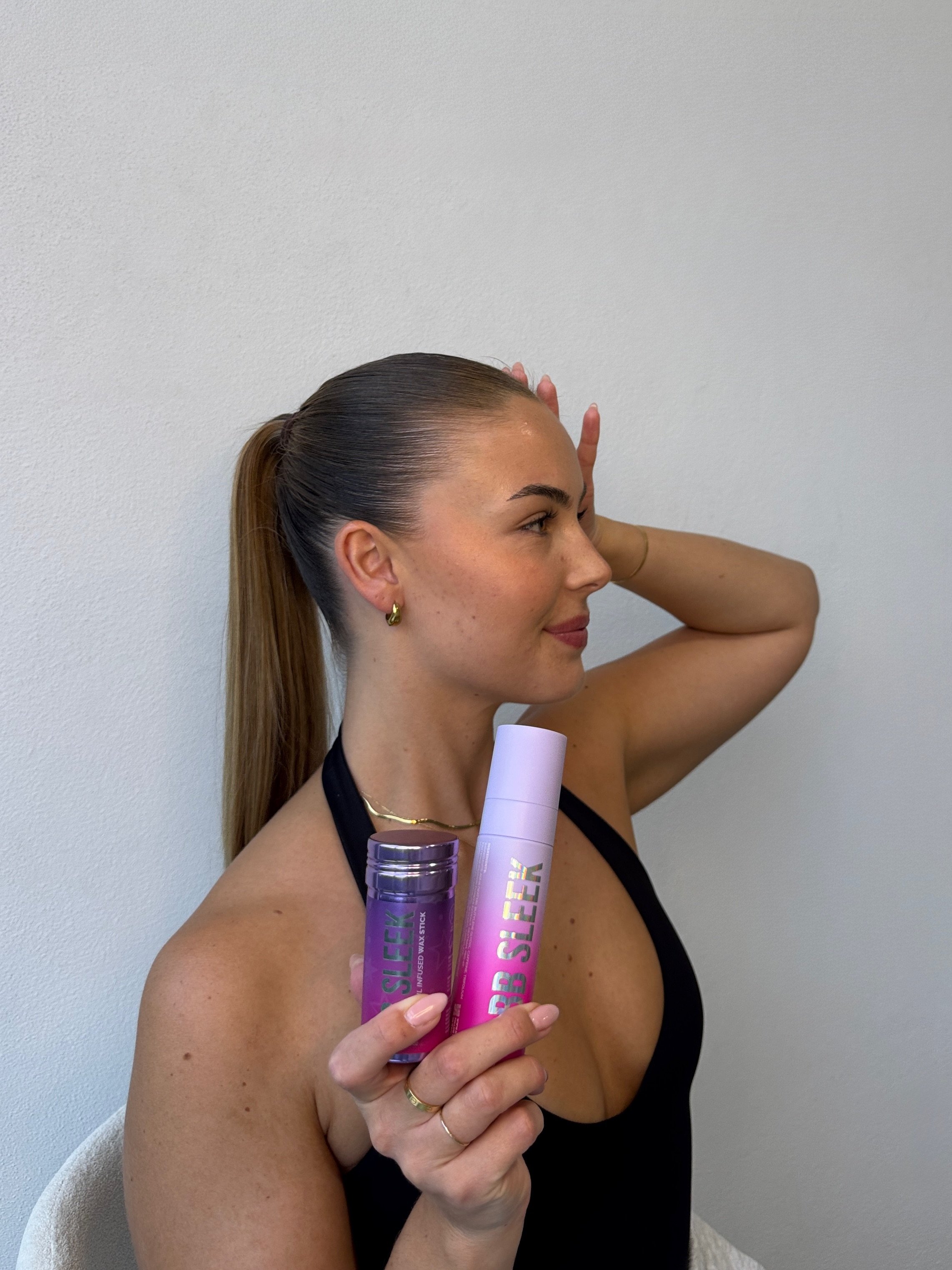 A woman with a sleek ponytail poses while holding a beauty product in one hand and a skincare bottle in the other, showcasing a confident expression.