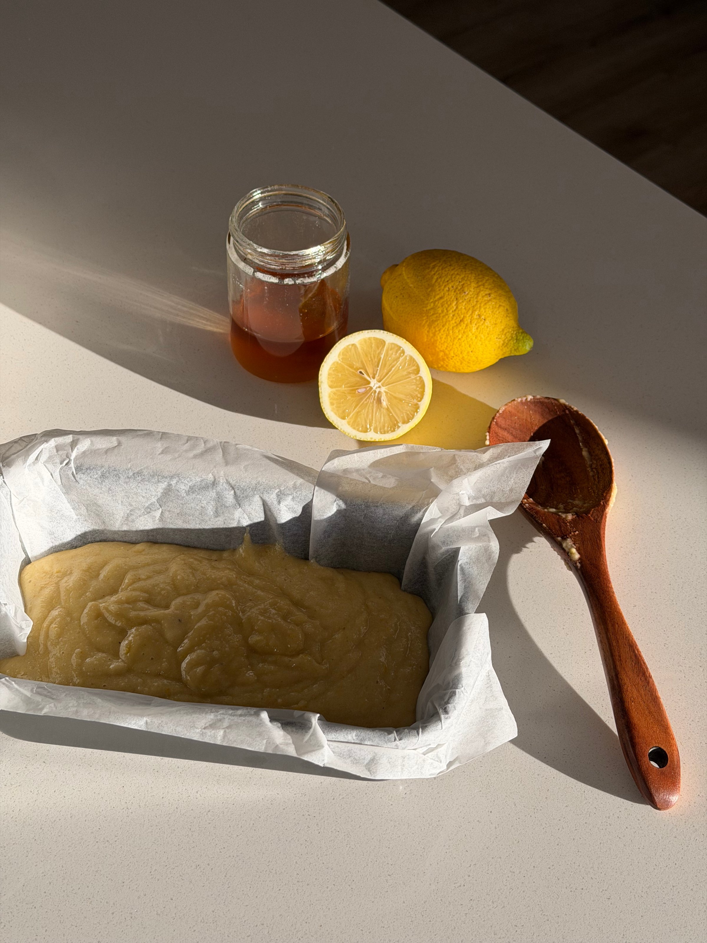 A loaf of lemon batter in a parchment-lined baking dish, accompanied by a jar of honey, a whole lemon, and a halved lemon on a light-colored countertop.