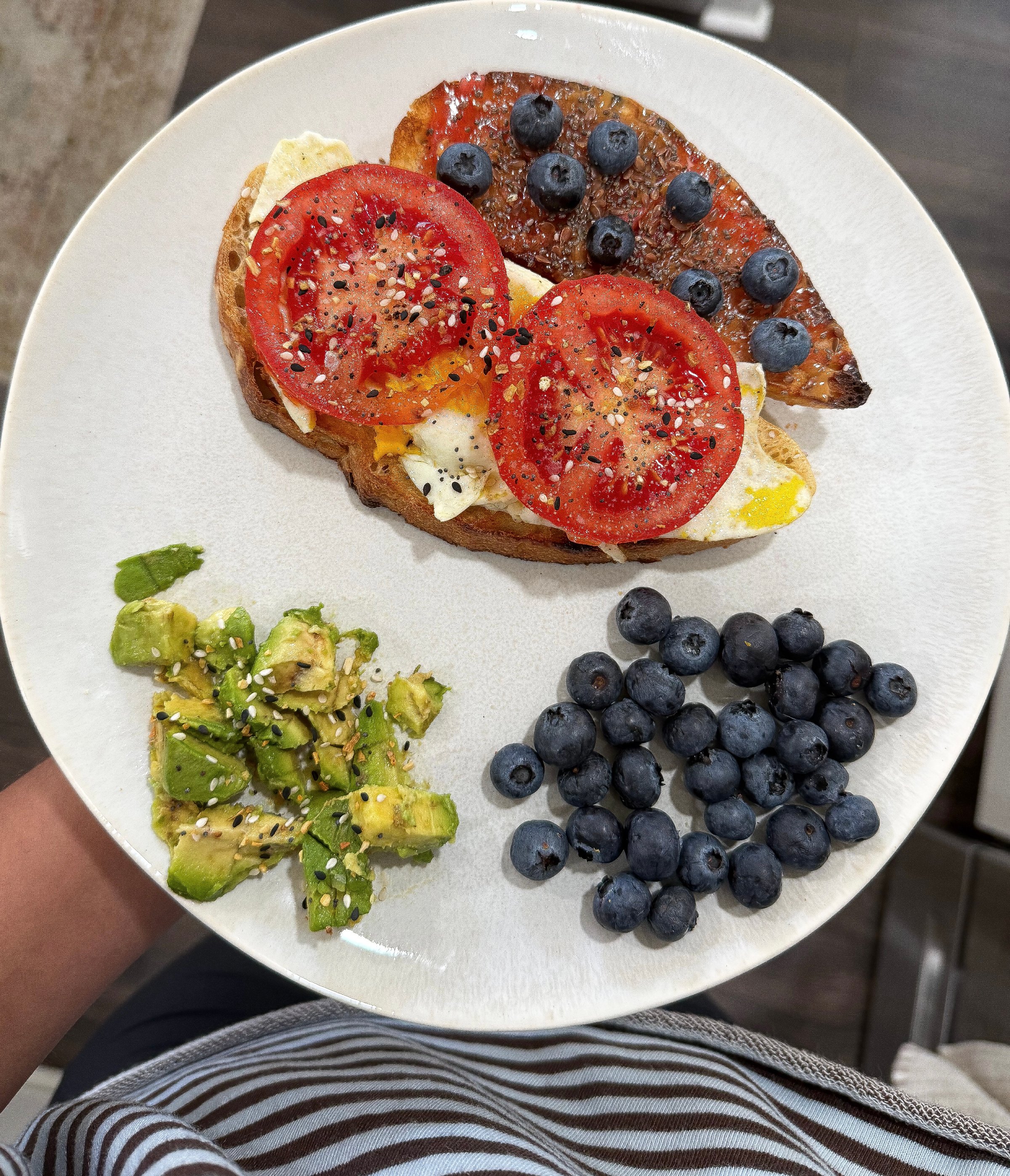 A plate featuring a slice of toast topped with sliced tomatoes, blueberries, and a side of avocado salad, showcasing a colorful and healthy meal.