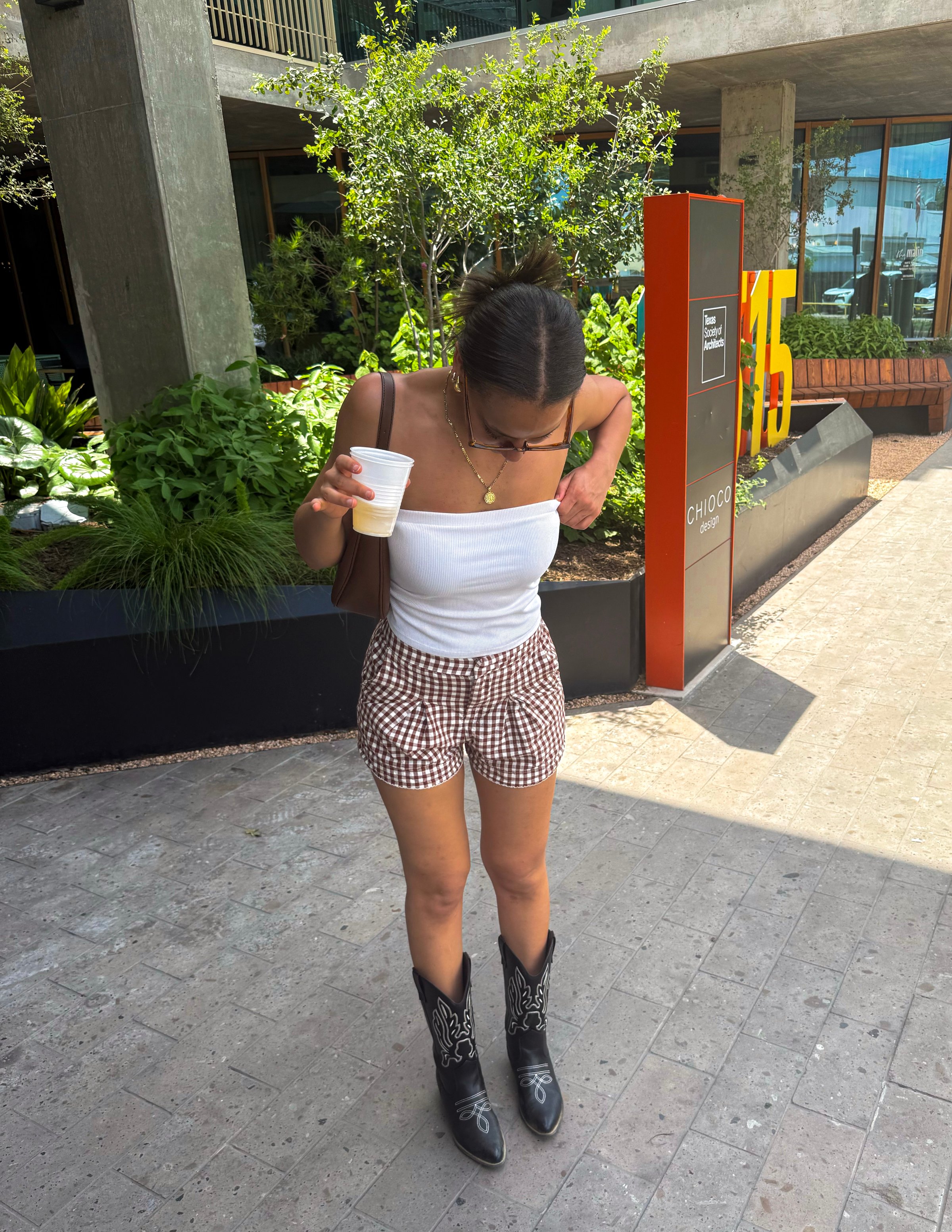 A young woman stands outdoors, wearing a white strapless top and checkered shorts, holding a drink. She is wearing black cowboy boots and looking down at her outfit.