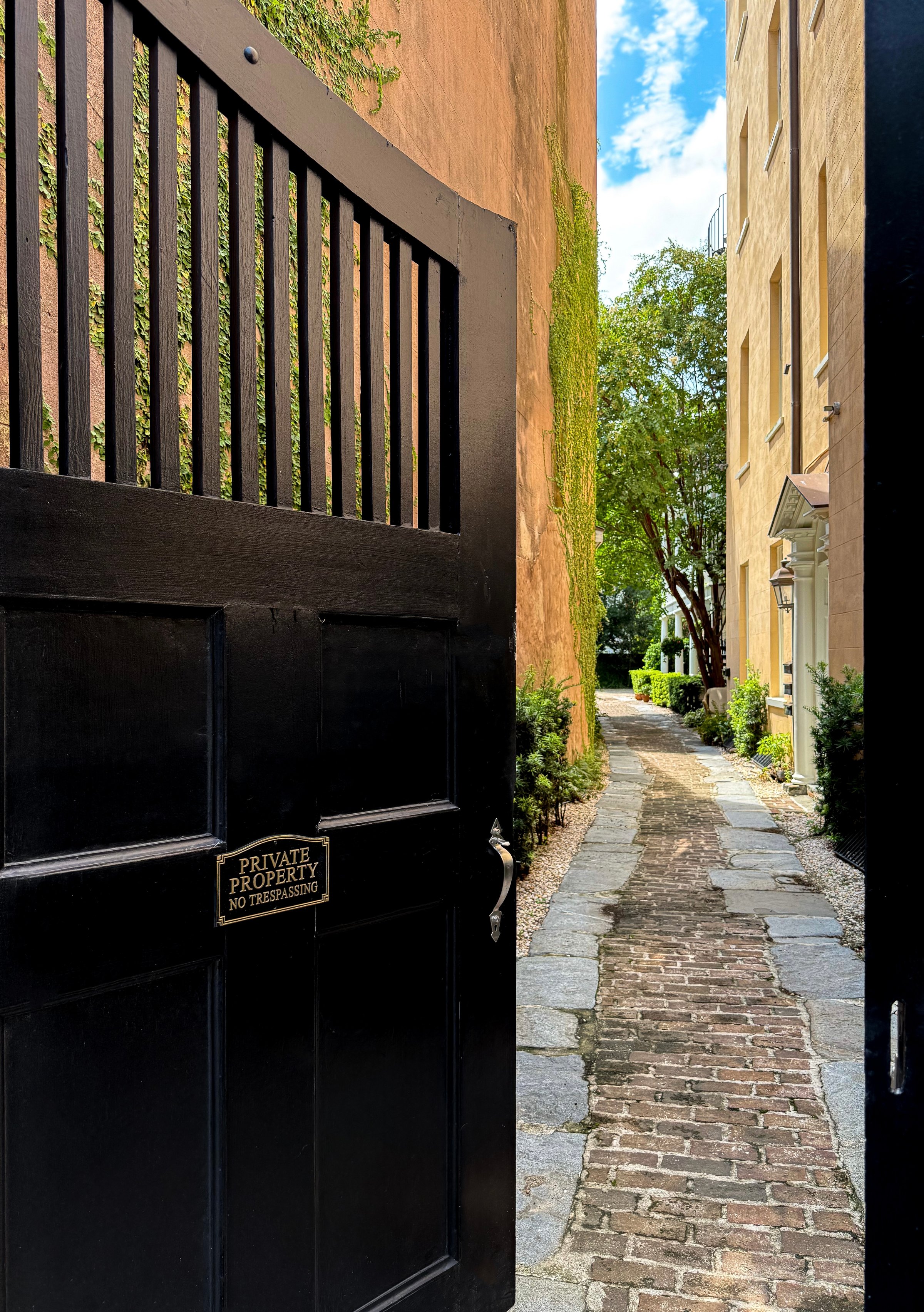 A black gate partially open, revealing a cobblestone pathway lined with greenery and buildings under a blue sky.