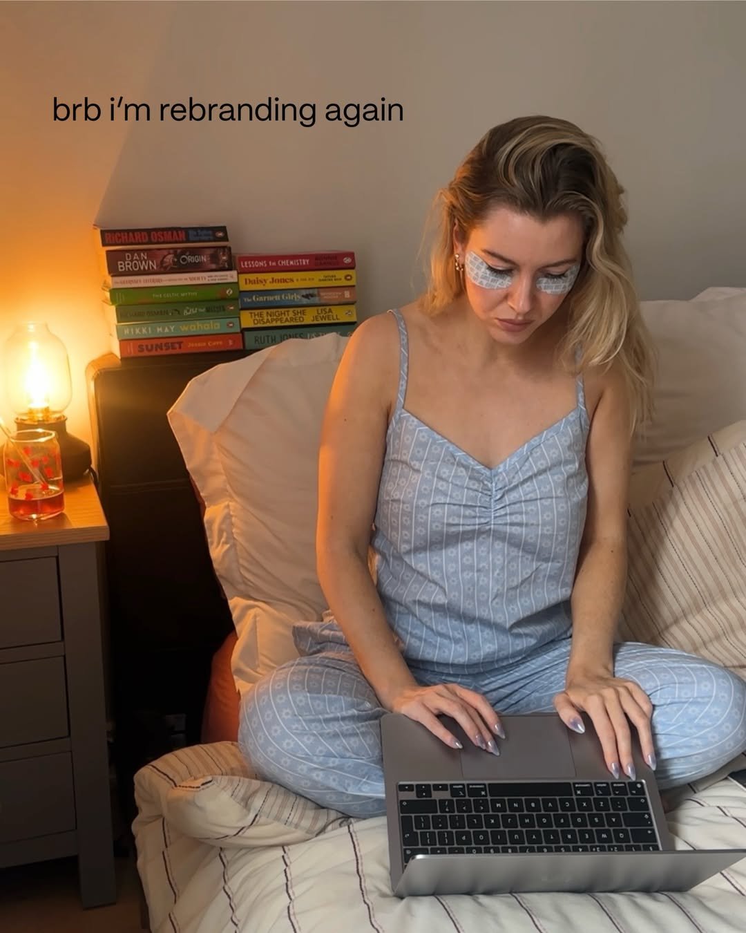 A person sitting on a bed in pajamas, working on a laptop with colorful books in the background. The scene is cozy and casual, with a warm lamp illuminating the space.