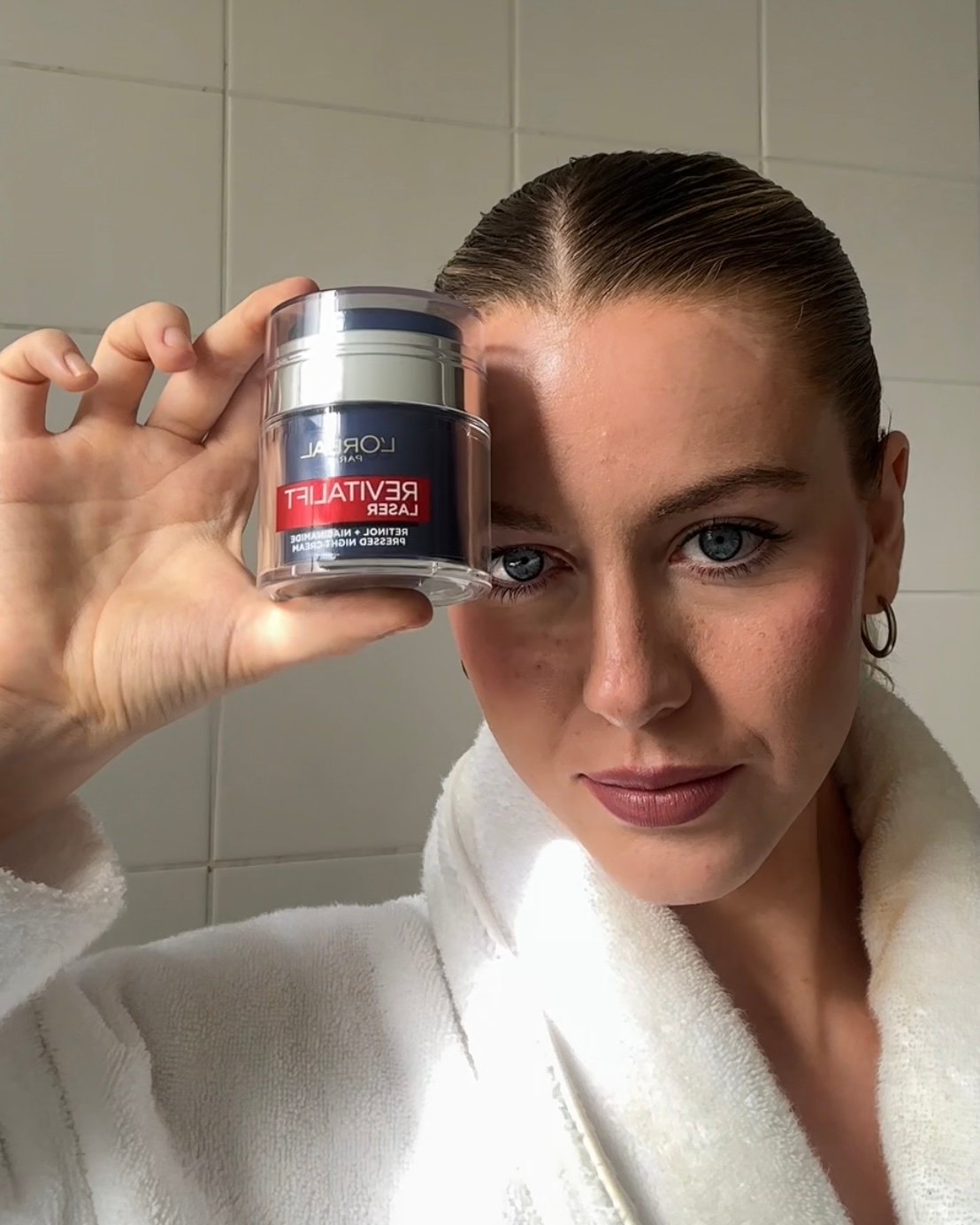 A young woman in a white robe holds a jar of L'Oreal Revitalift skincare product close to her face, showcasing her clear skin and natural makeup.