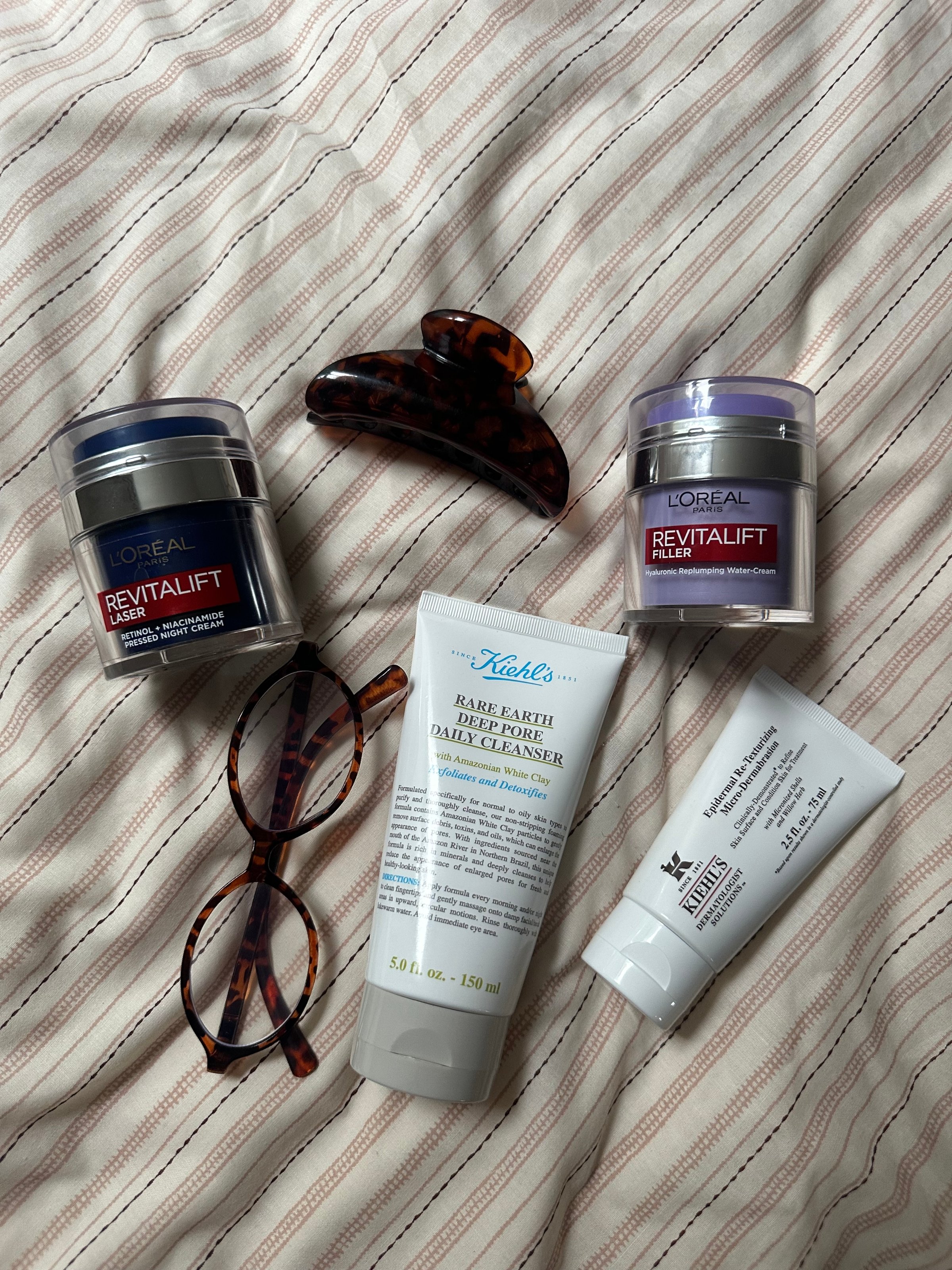 A flat lay arrangement of skincare products including L'Oreal Revitalift creams, a Kiehl's cleanser, and a small tube of moisturizer, alongside a tortoiseshell hair clip and a pair of eyeglasses.