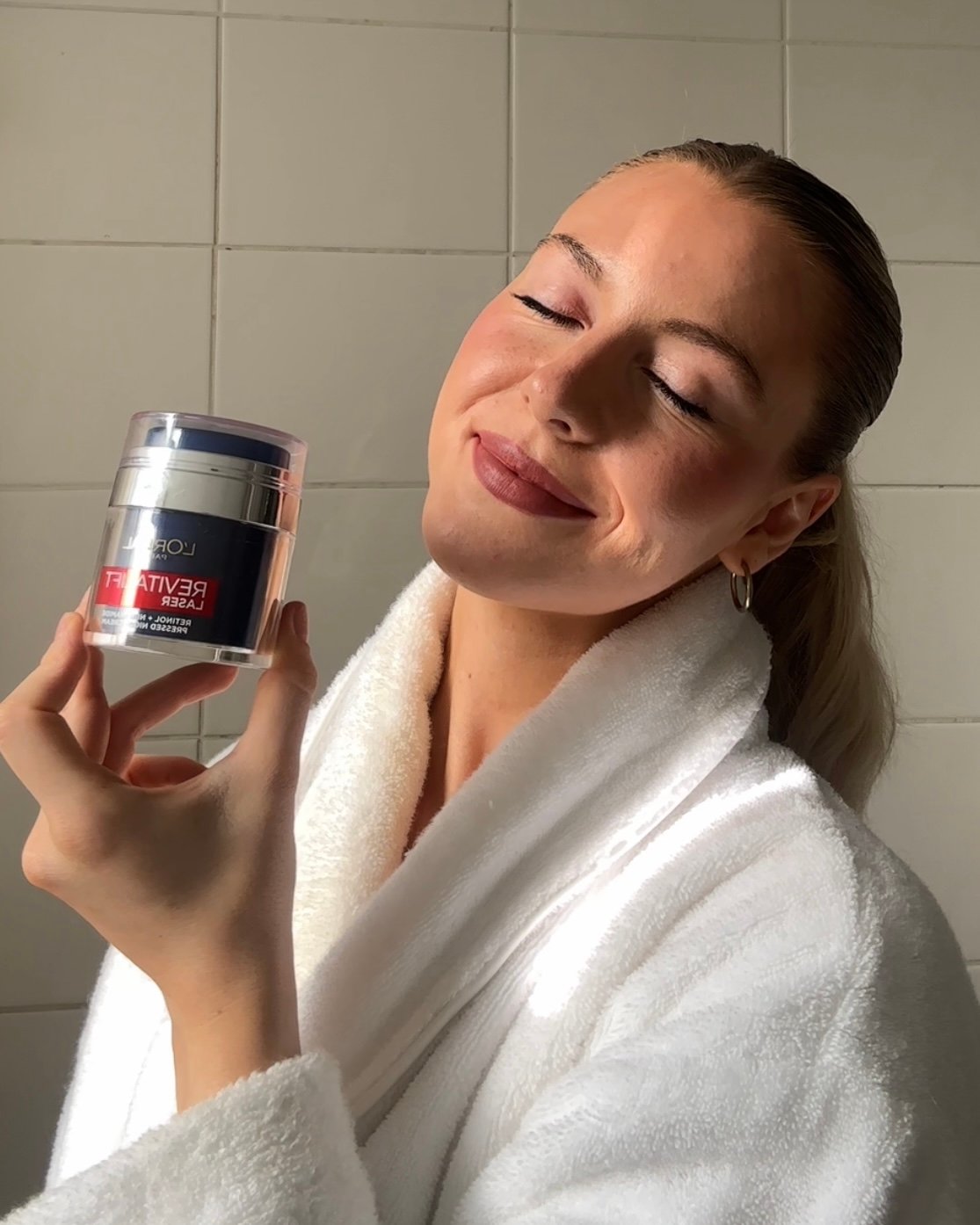 A woman in a white robe smiles while holding a skincare product in a bathroom setting.