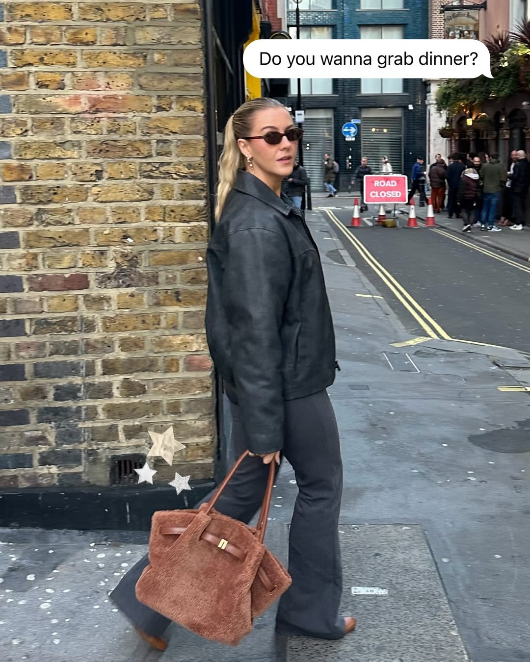 A woman in a black leather jacket and sunglasses stands against a brick wall, holding a brown handbag. She appears to be in an urban setting, with people and shops in the background.