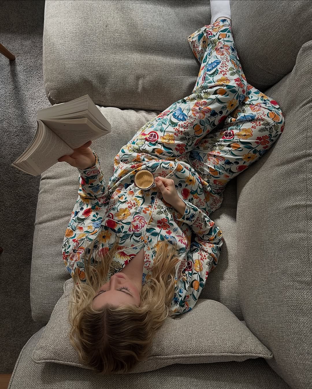 A person lounging on a couch, wearing floral pajamas and reading a book while holding a small cup.