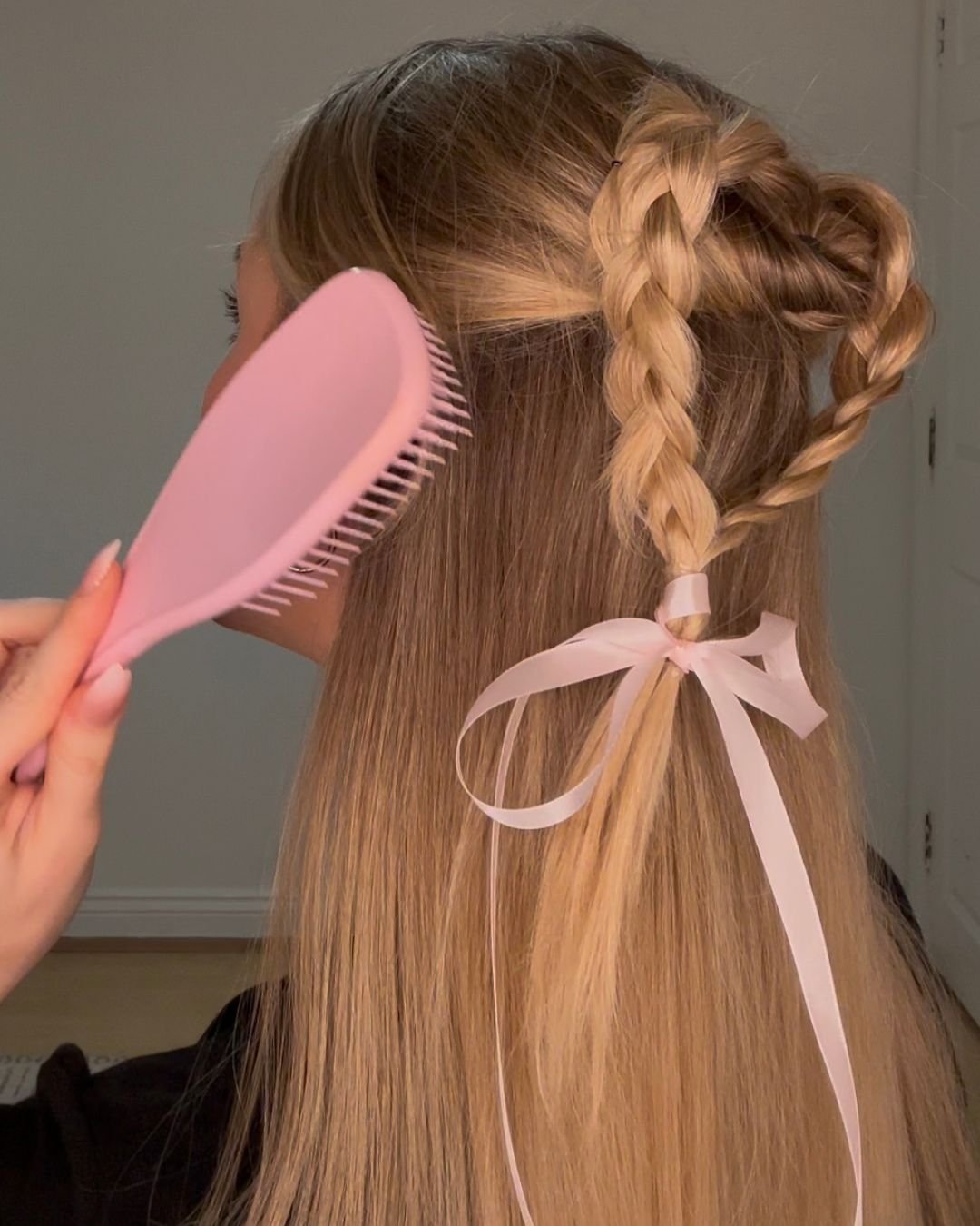 A person styling their hair with a pink comb, showcasing a braided hairstyle adorned with a ribbon.