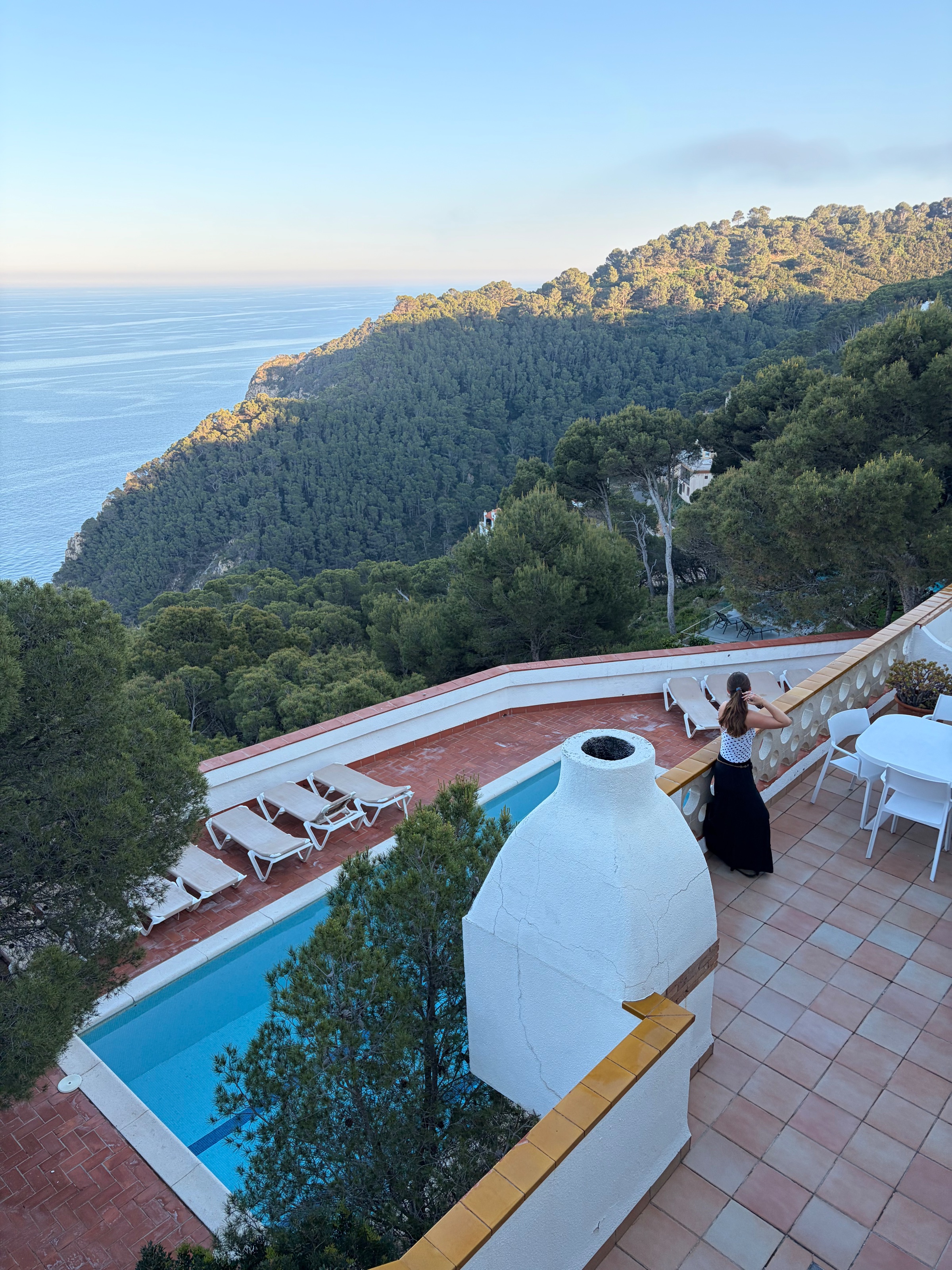 A woman in a black dress stands by a serene pool overlooking a mountainous landscape and the sea in the distance.