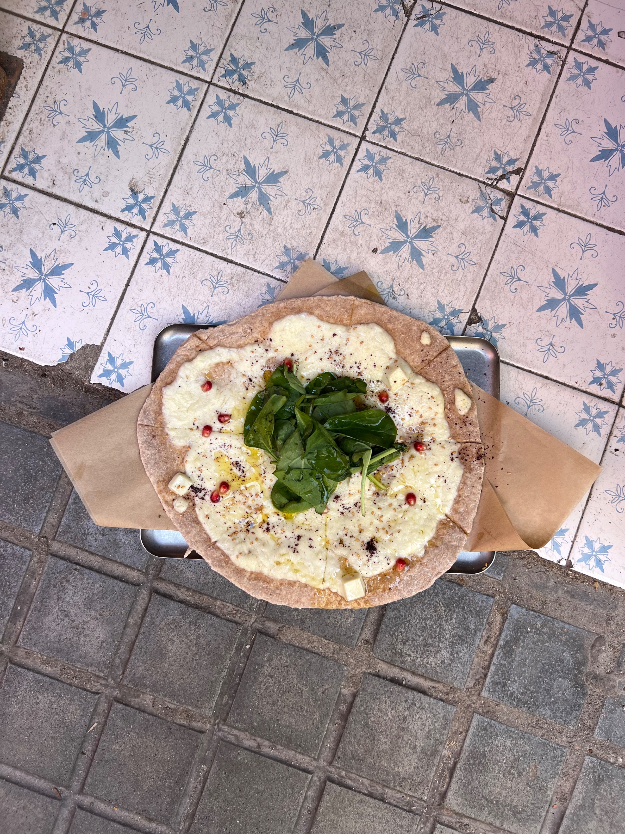 A flatbread topped with cheese, spinach, and pomegranate seeds, presented on a rustic tray against a patterned tiled background.