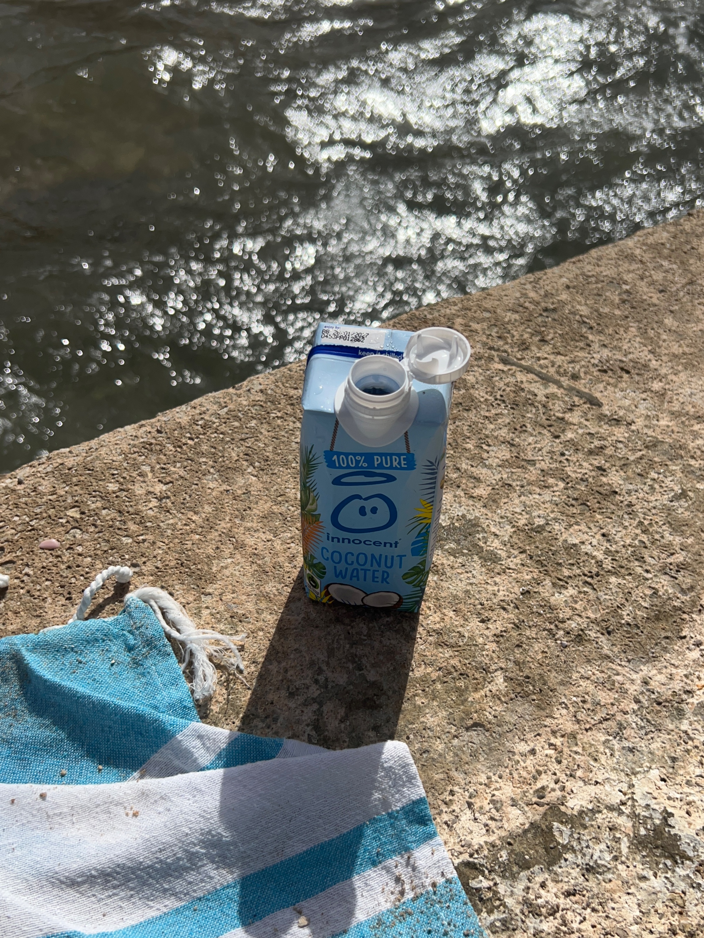 A coconut water carton placed on a stone surface near a body of water, with a beach towel partially visible.
