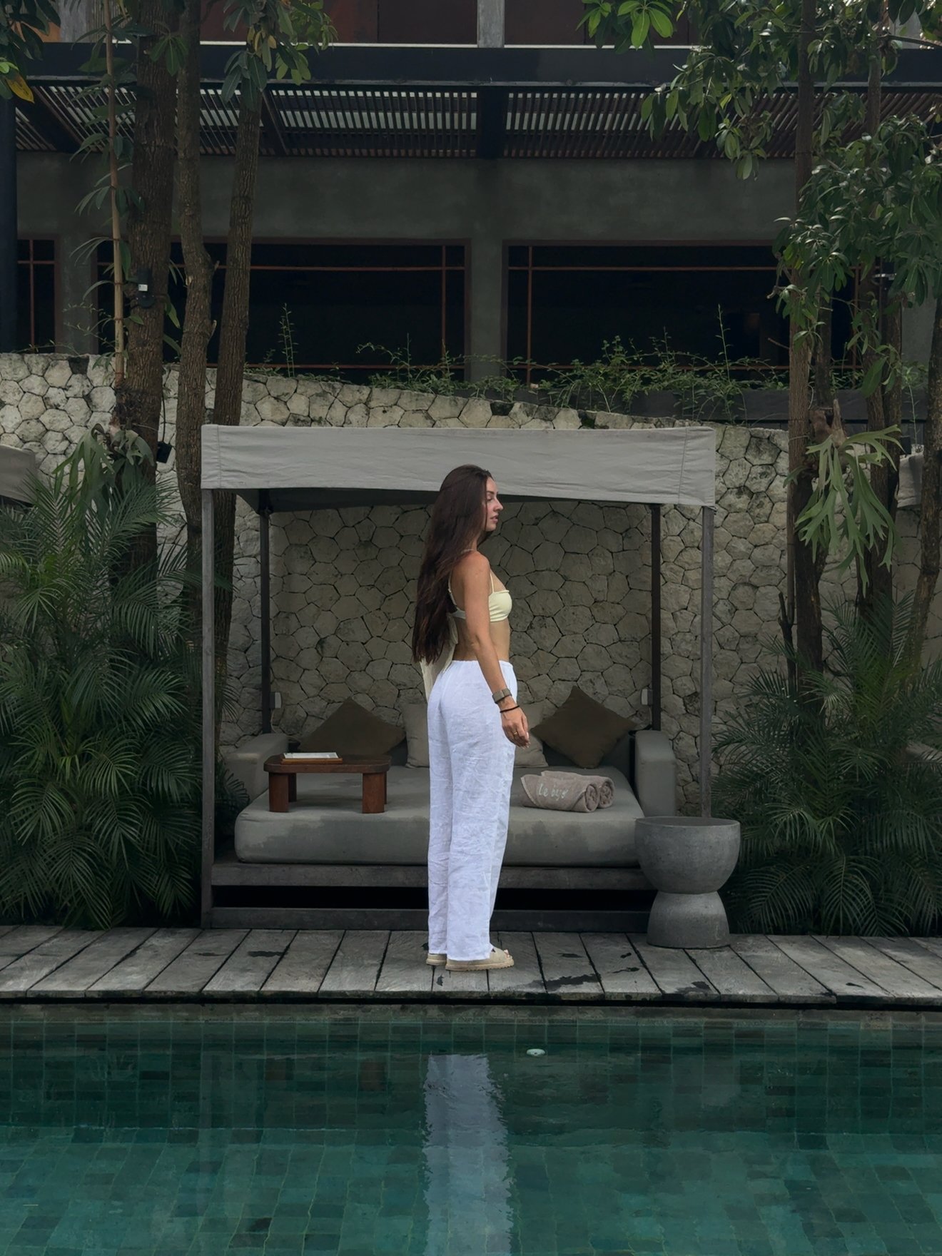 A woman stands by a poolside cabana in a tropical setting, wearing a stylish outfit and looking away from the camera.