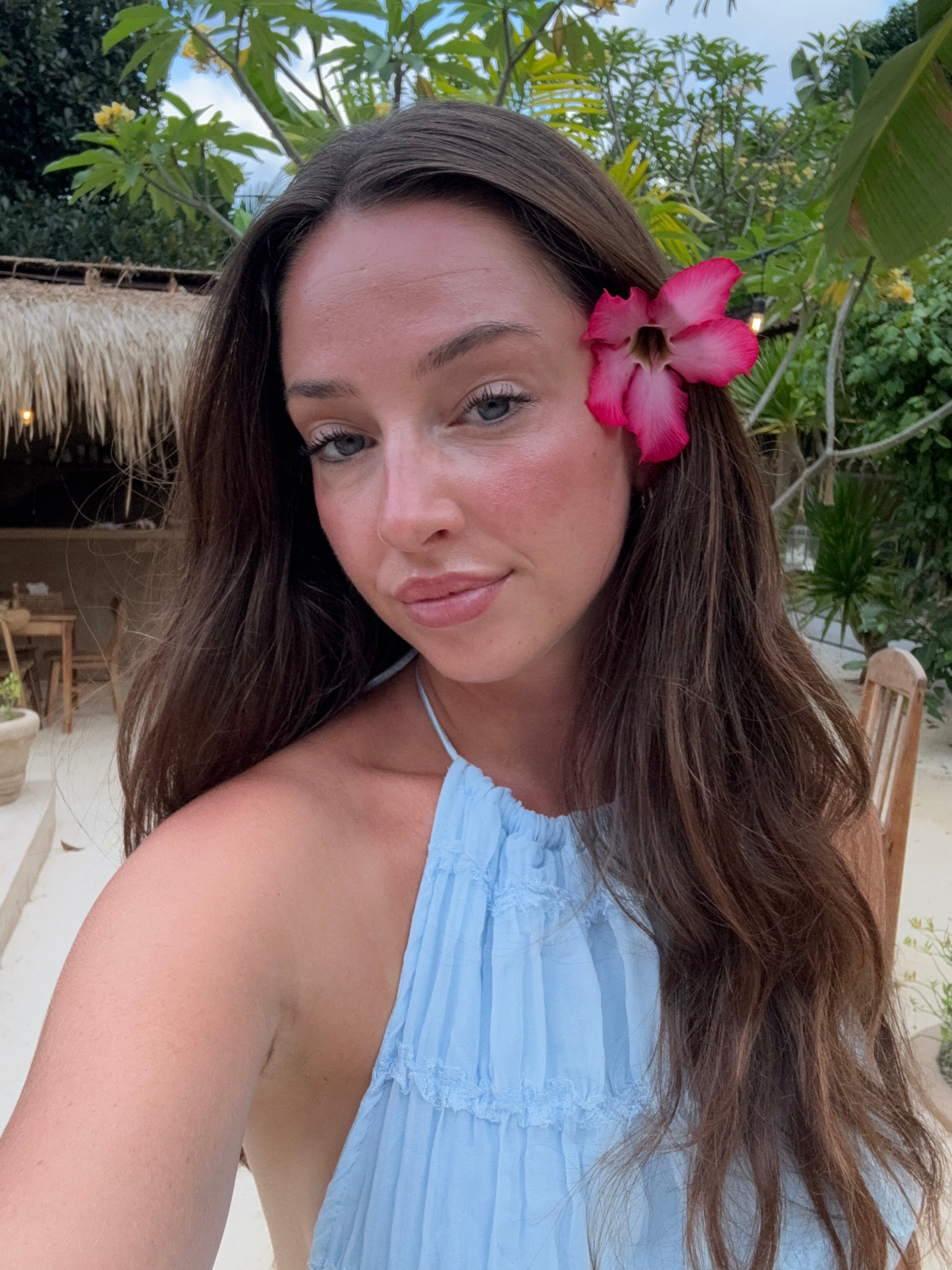 A woman with long brown hair poses outdoors, wearing a light blue halter top and a pink flower tucked behind her ear, with a tropical background.