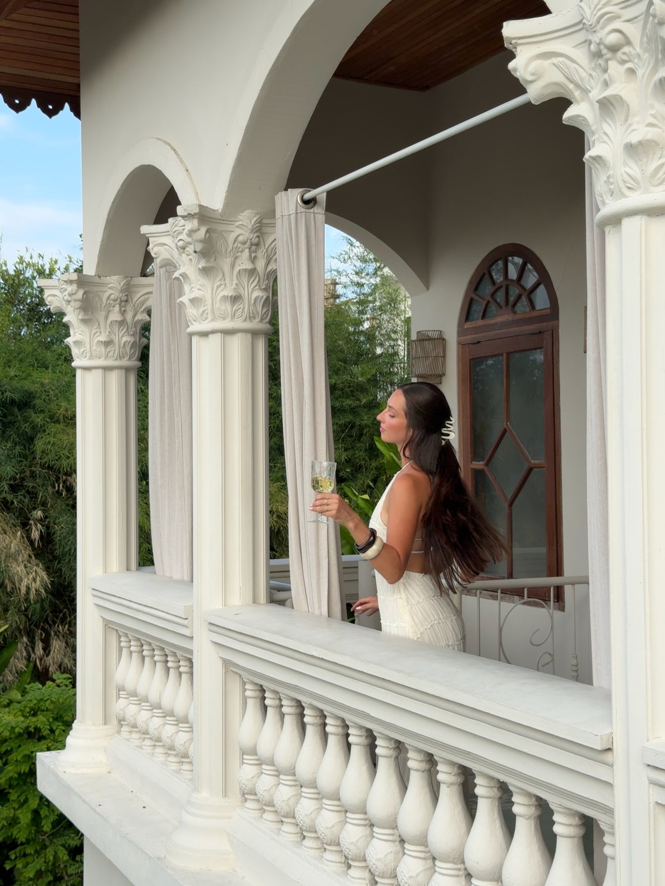 A woman in a wedding dress stands on a balcony, holding a drink and gazing thoughtfully into the distance, framed by elegant architectural columns.
