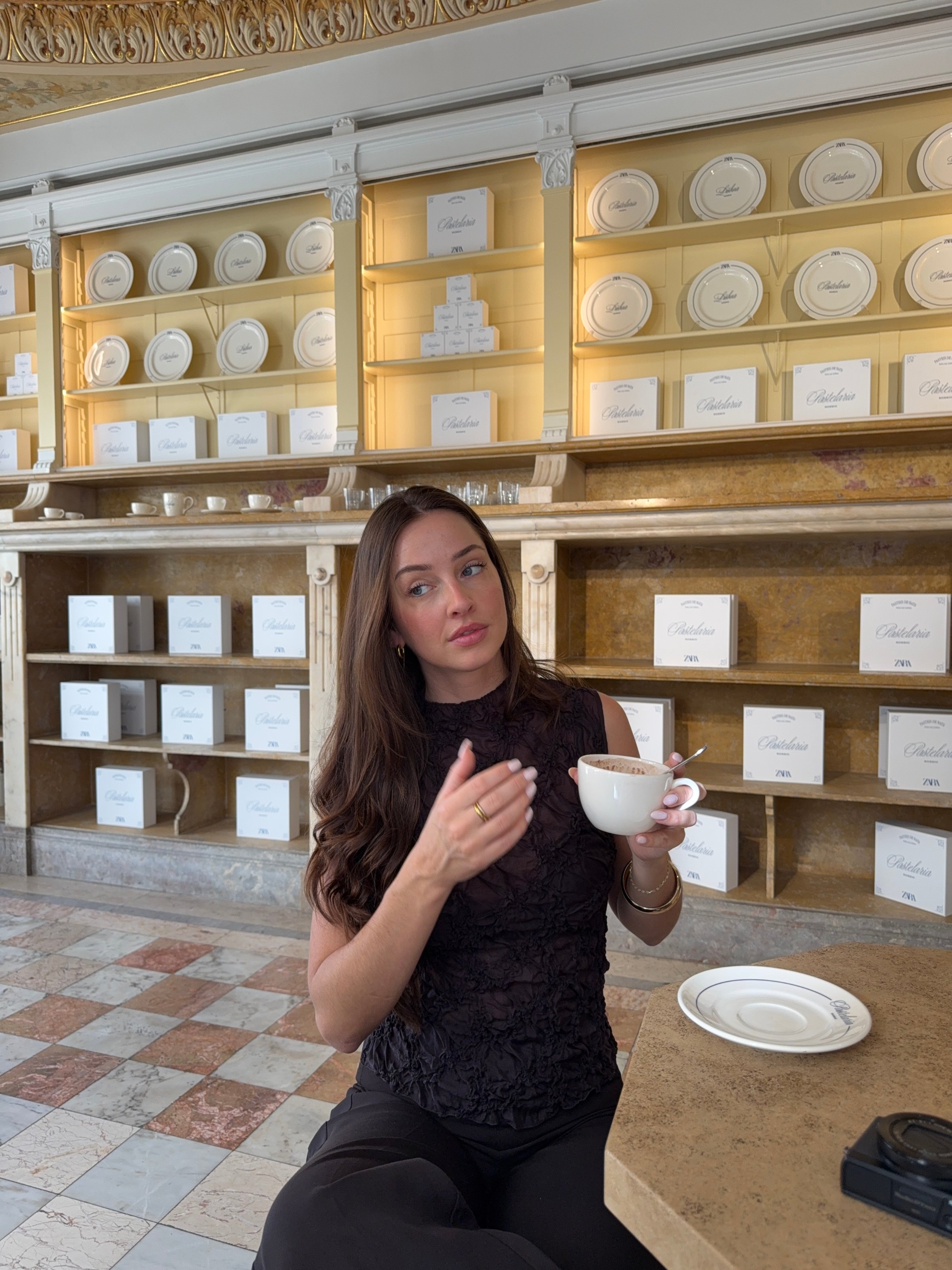 A woman sitting at a table in an elegant setting, holding a cup and gesturing while looking thoughtfully at the camera.