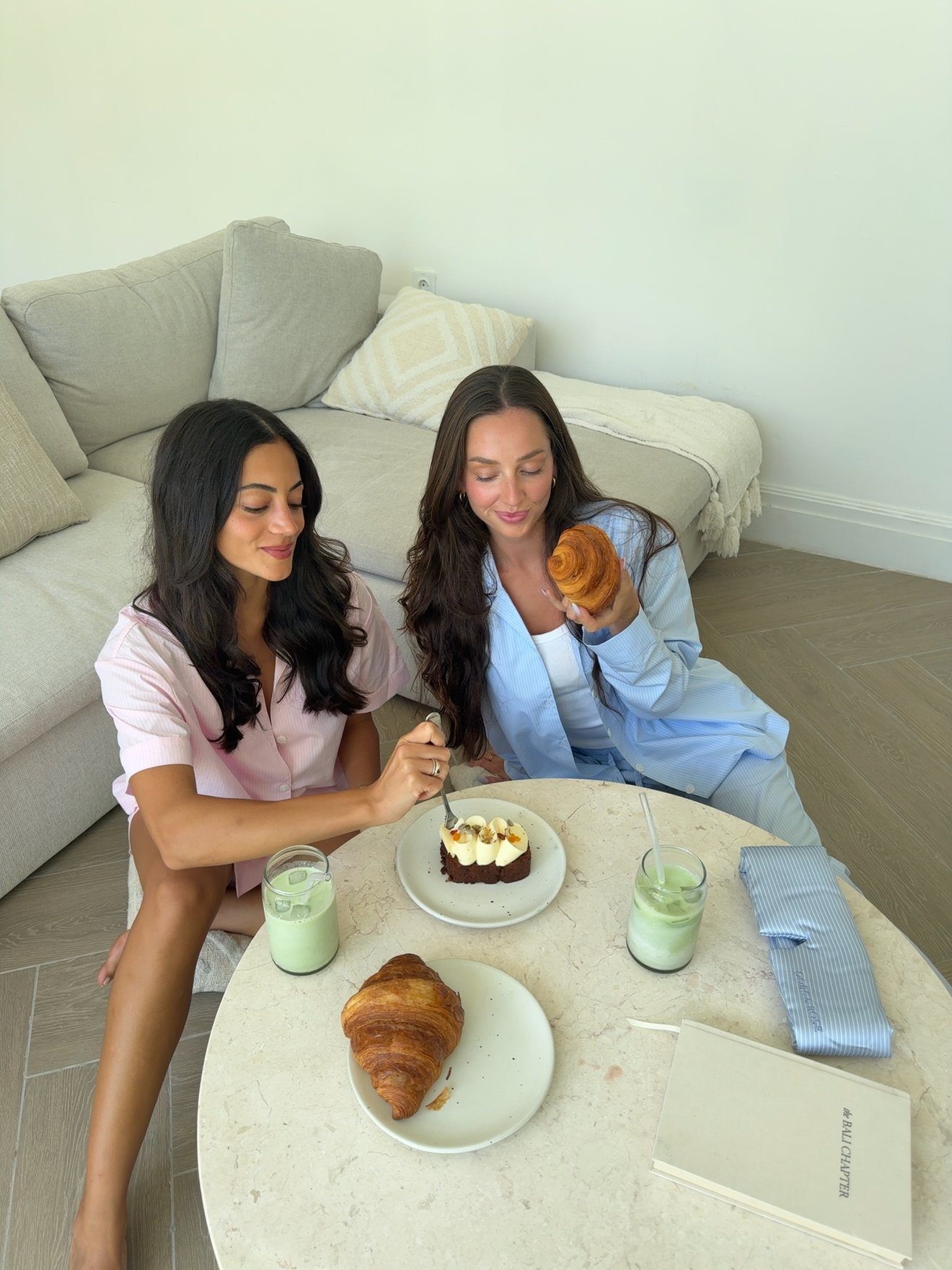 Two women enjoying pastries and drinks while seated on a couch in a bright, modern living space.