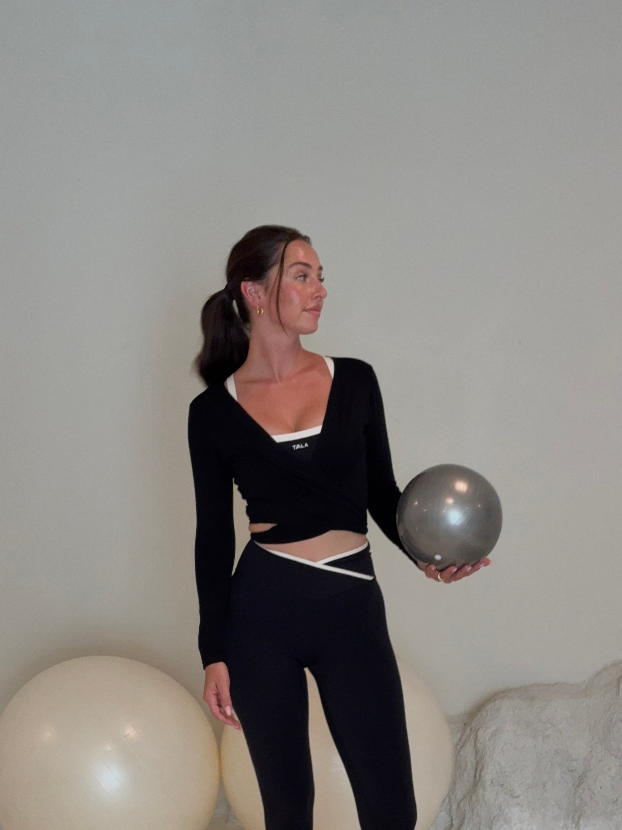 A woman in a black athletic outfit holds a silver exercise ball while standing in a minimalist studio with large exercise balls in the background.
