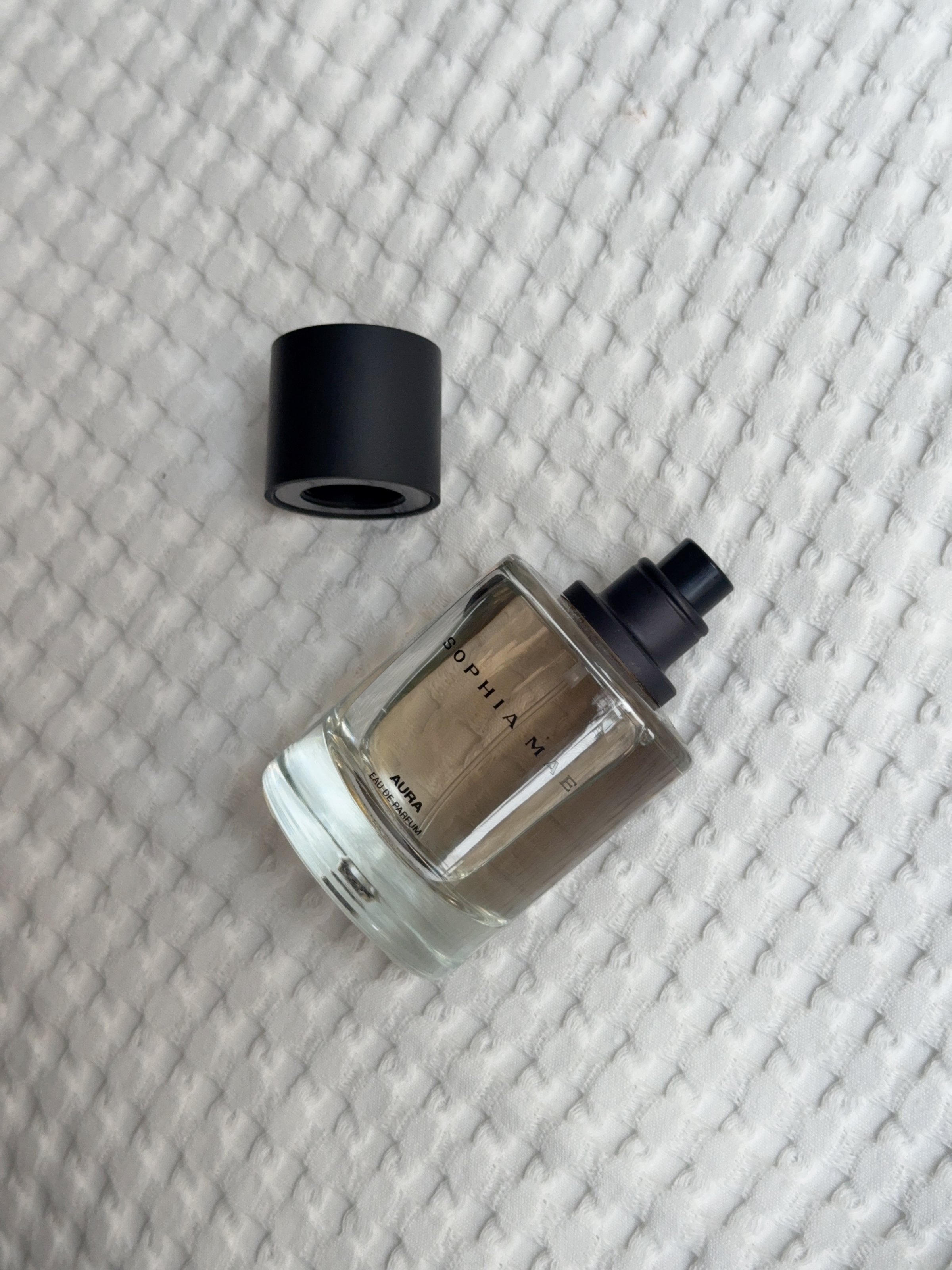 A glass perfume bottle with a black cap resting on a textured white surface.