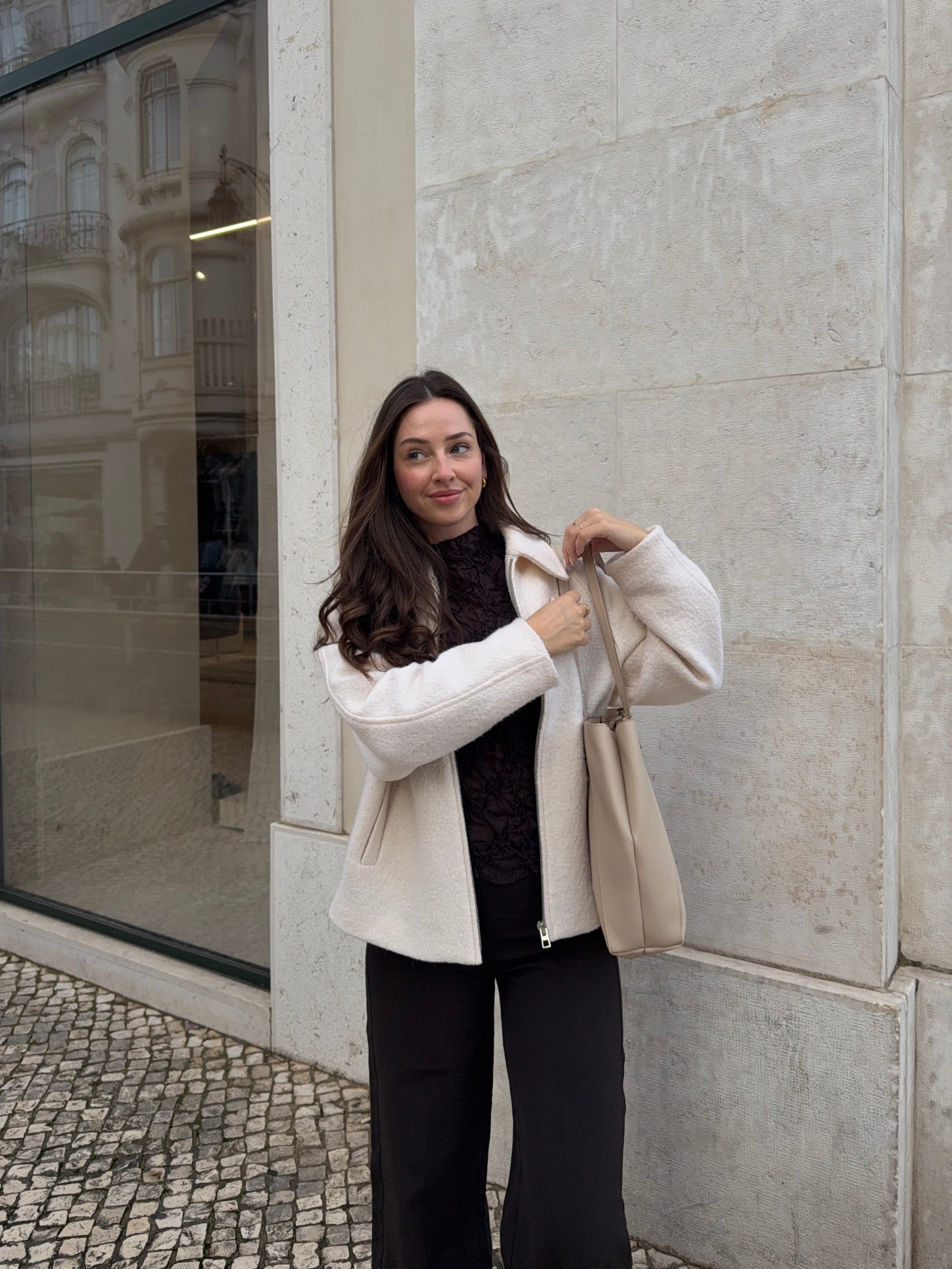 A woman stands outside a modern building, wearing a cozy white jacket and black pants, holding a beige bag.