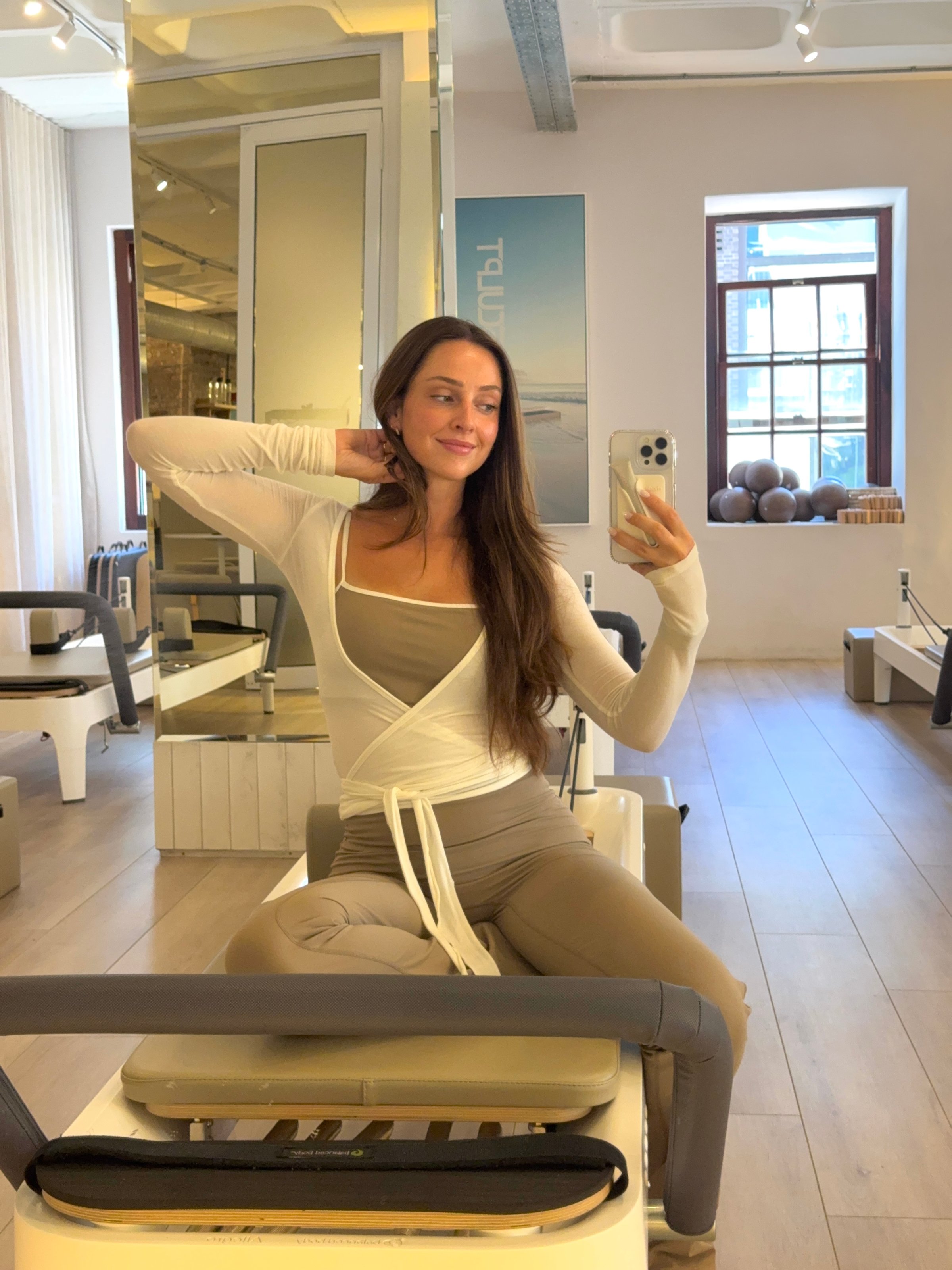 A woman poses for a selfie in a modern fitness studio, wearing stylish activewear and smiling at the camera.