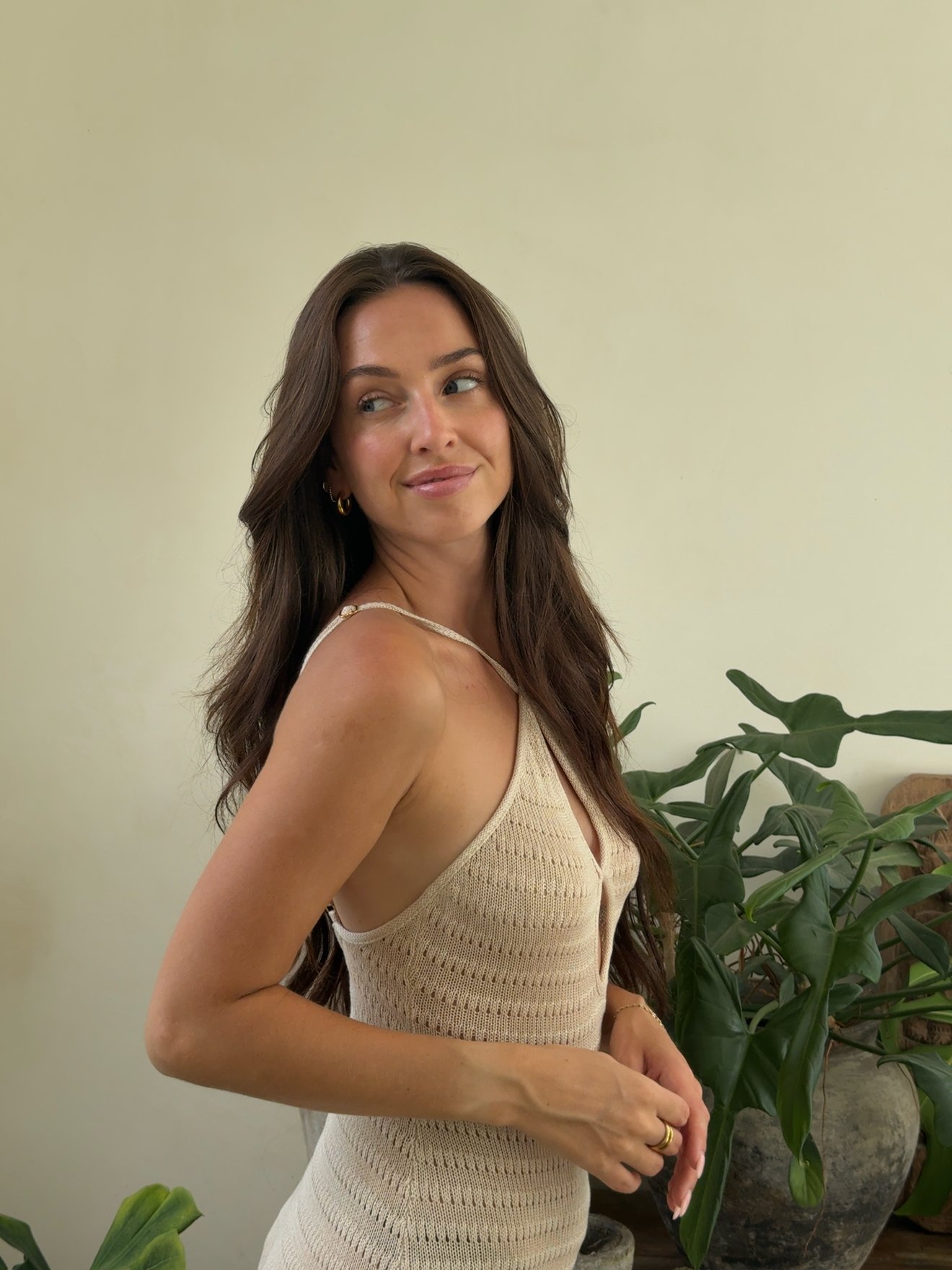 A woman with long, wavy hair poses gracefully against a neutral background, showcasing a knitted beige top. Green plants are visible in the scene, adding a natural touch.