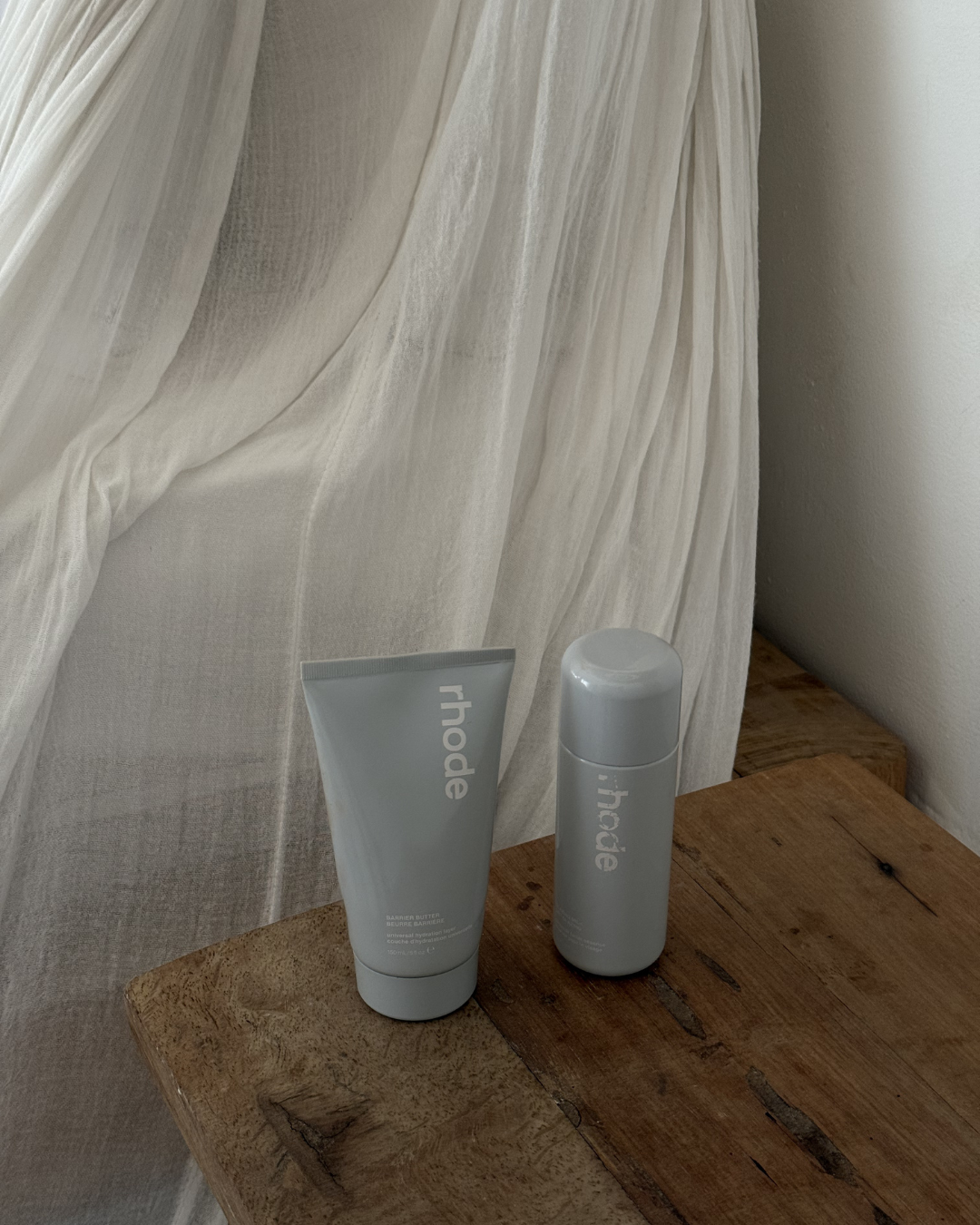 A minimalist display of skincare products on a wooden surface, with soft fabric draping in the background.