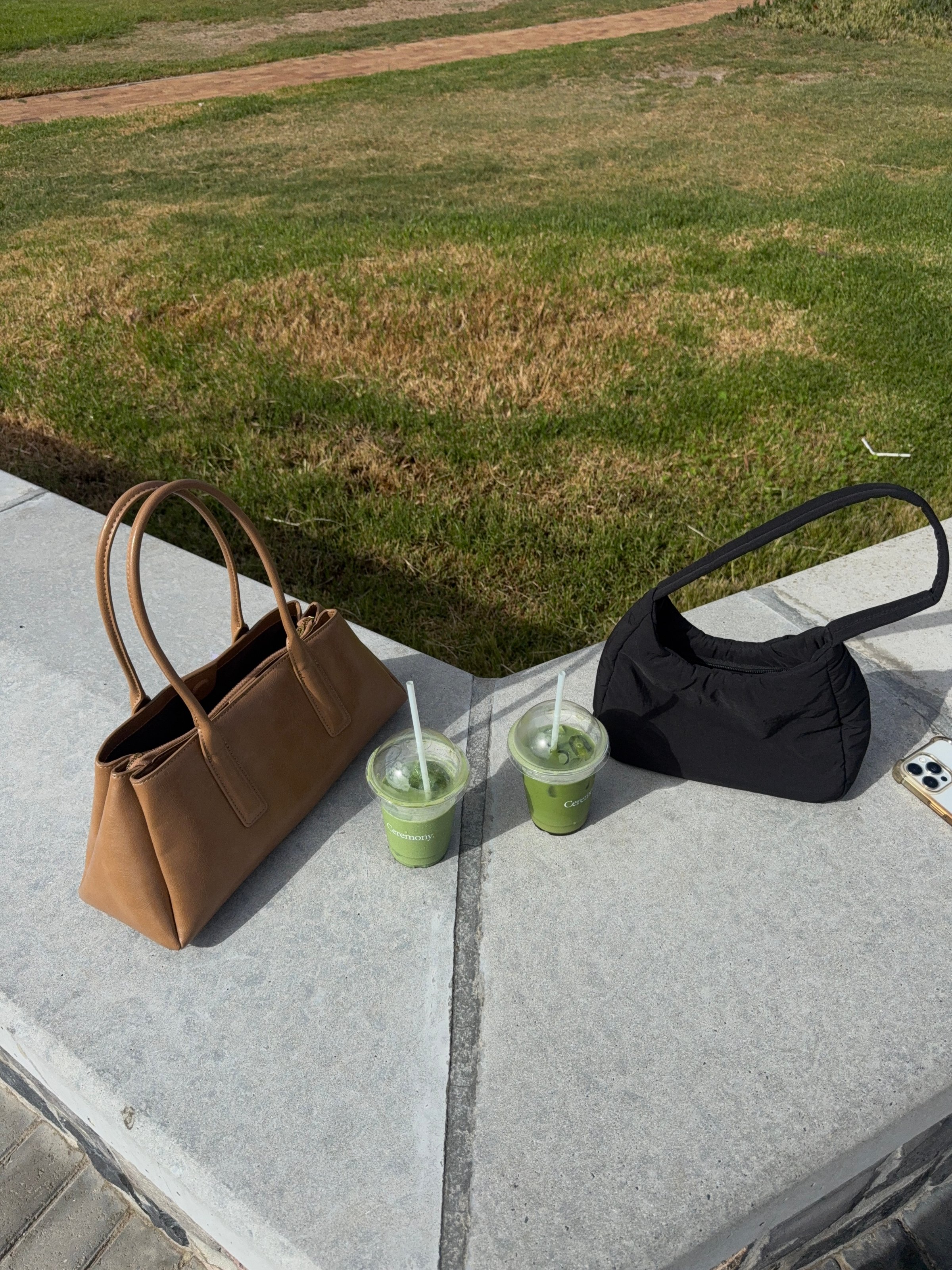 Two handbags placed on a stone surface next to two cups of green drinks, set against a grassy background.