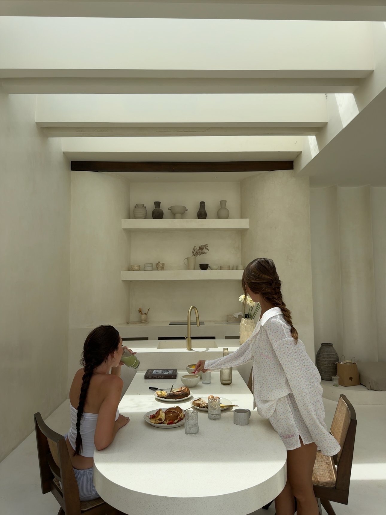 Two women enjoying breakfast at a modern, minimalist kitchen table with a variety of food items and drinks.