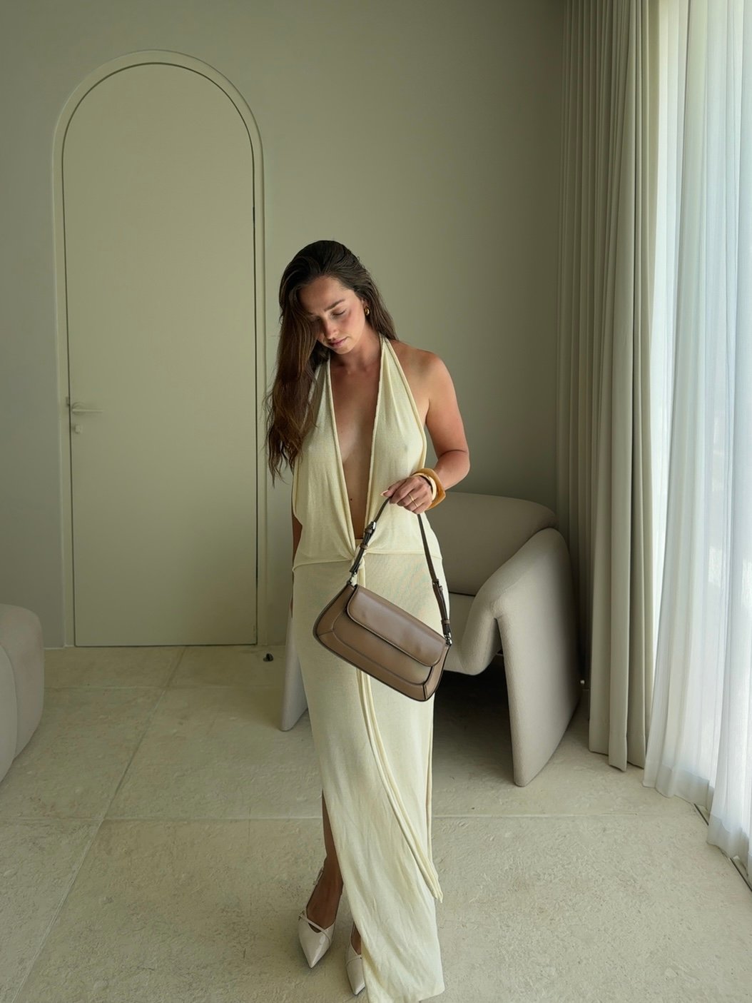 A woman in a stylish, light-colored dress stands in a modern interior, holding a handbag and looking down.