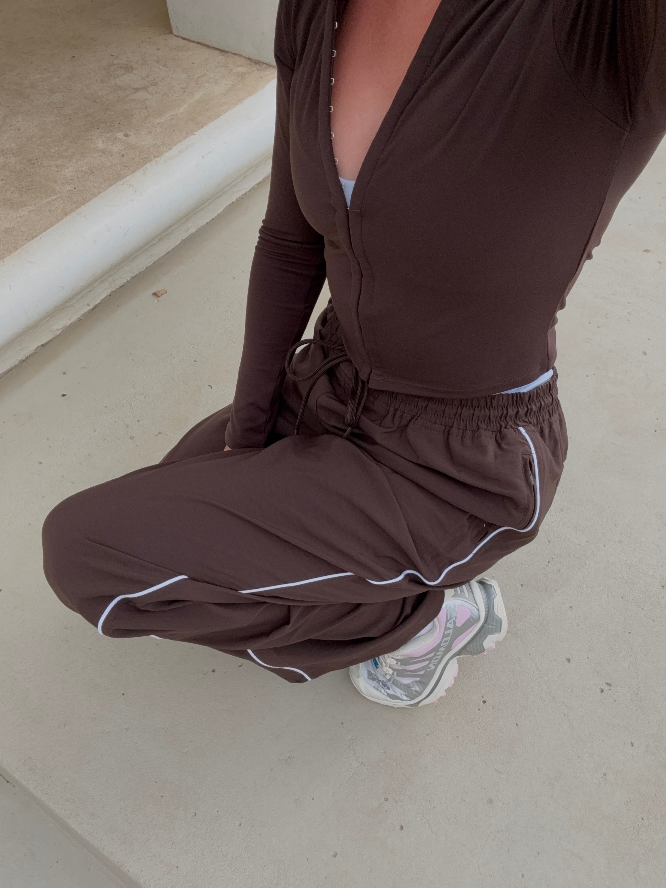 A person kneeling on a concrete surface, wearing a brown athletic outfit with white accents and sneakers.