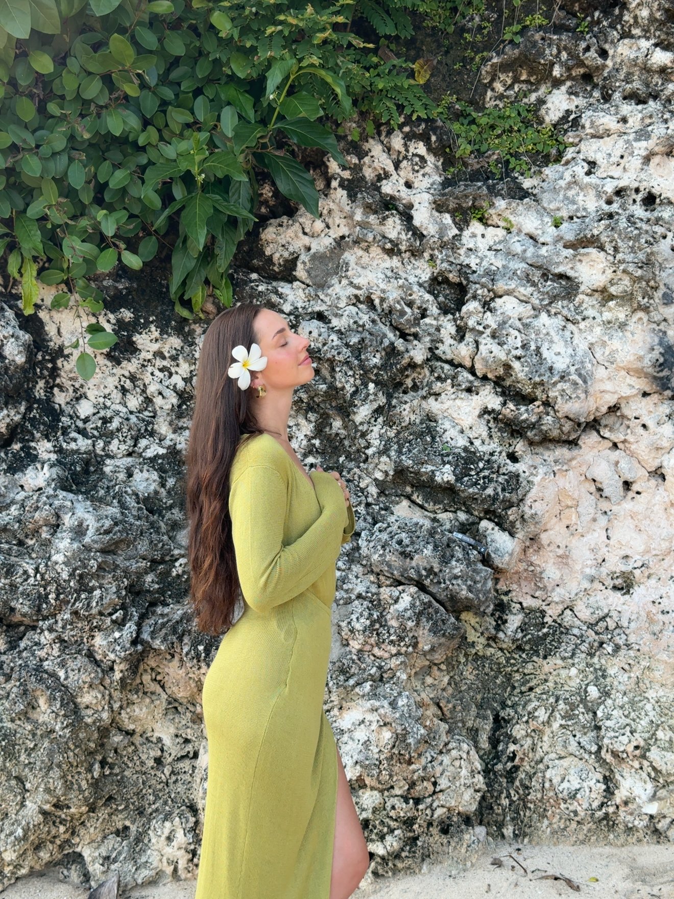A woman with long hair stands on a beach, wearing a green dress and a white flower in her hair, with rocky cliffs in the background.