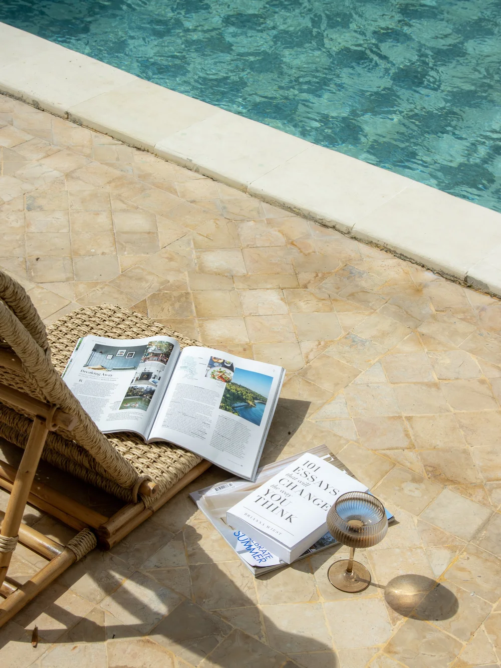 A relaxing outdoor scene featuring a woven lounge chair beside a pool, with an open magazine and a cocktail glass on the ground.