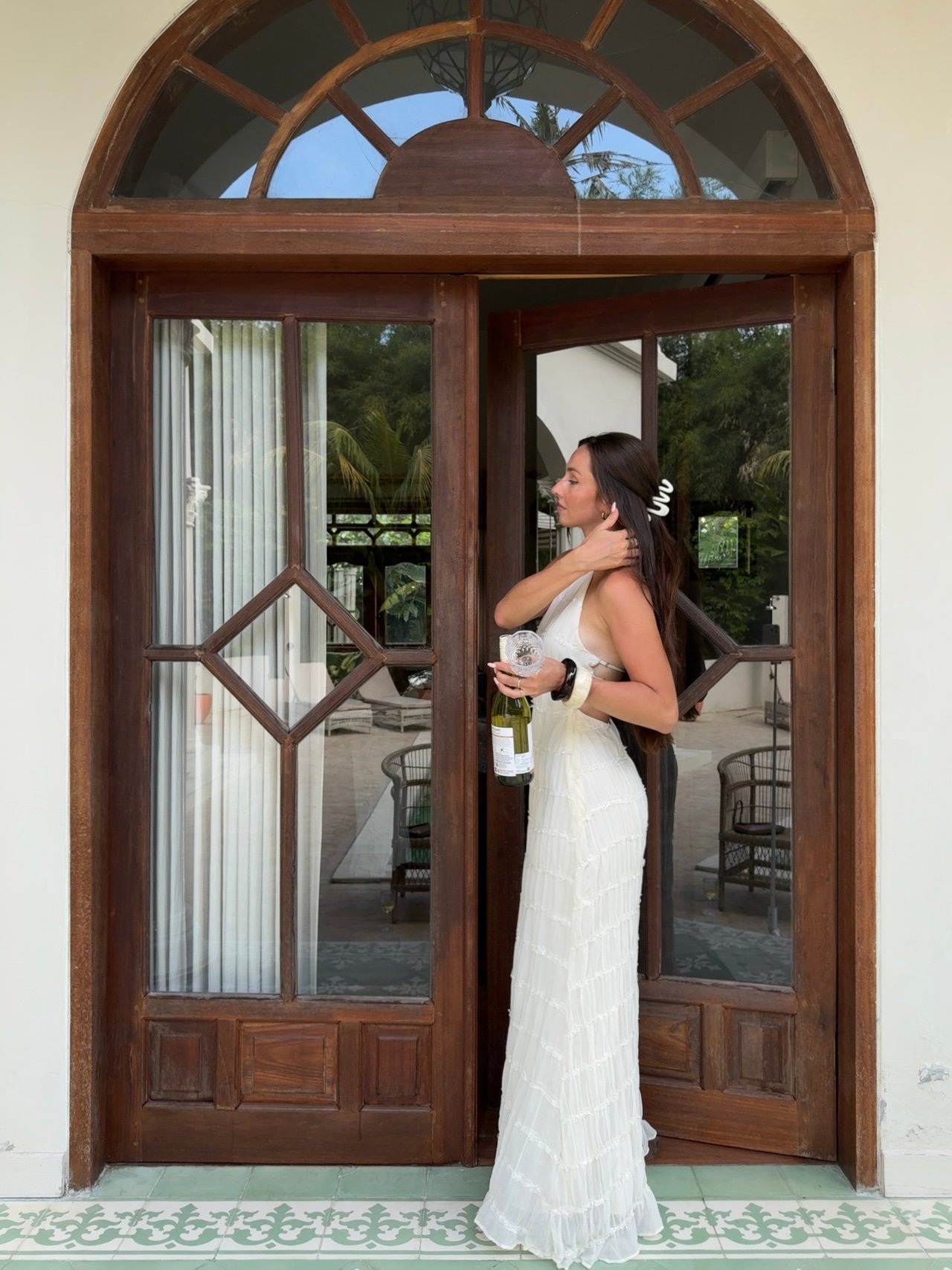 A woman in a white dress stands by a wooden door, holding a drink and looking away. The background features a glimpse of a stylish interior.