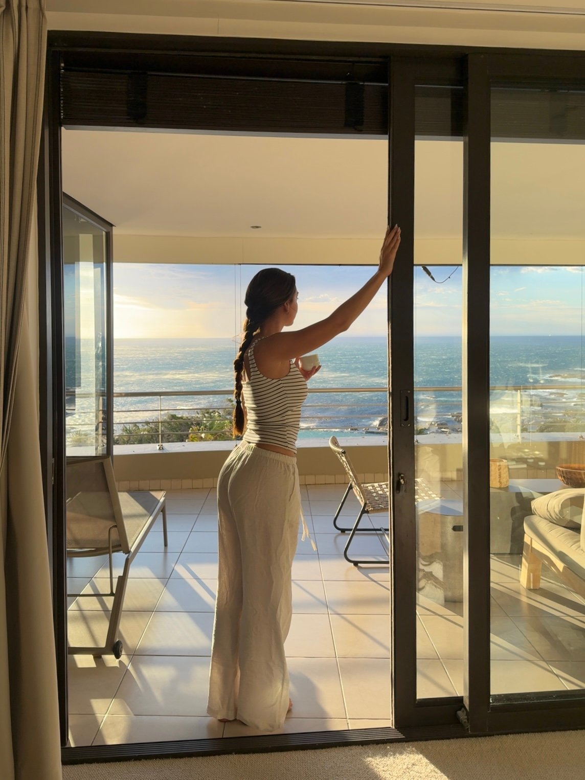 A woman stands by a large glass door, looking out at a scenic ocean view during sunset. She wears a striped top and white pants, with her hair in a braid.