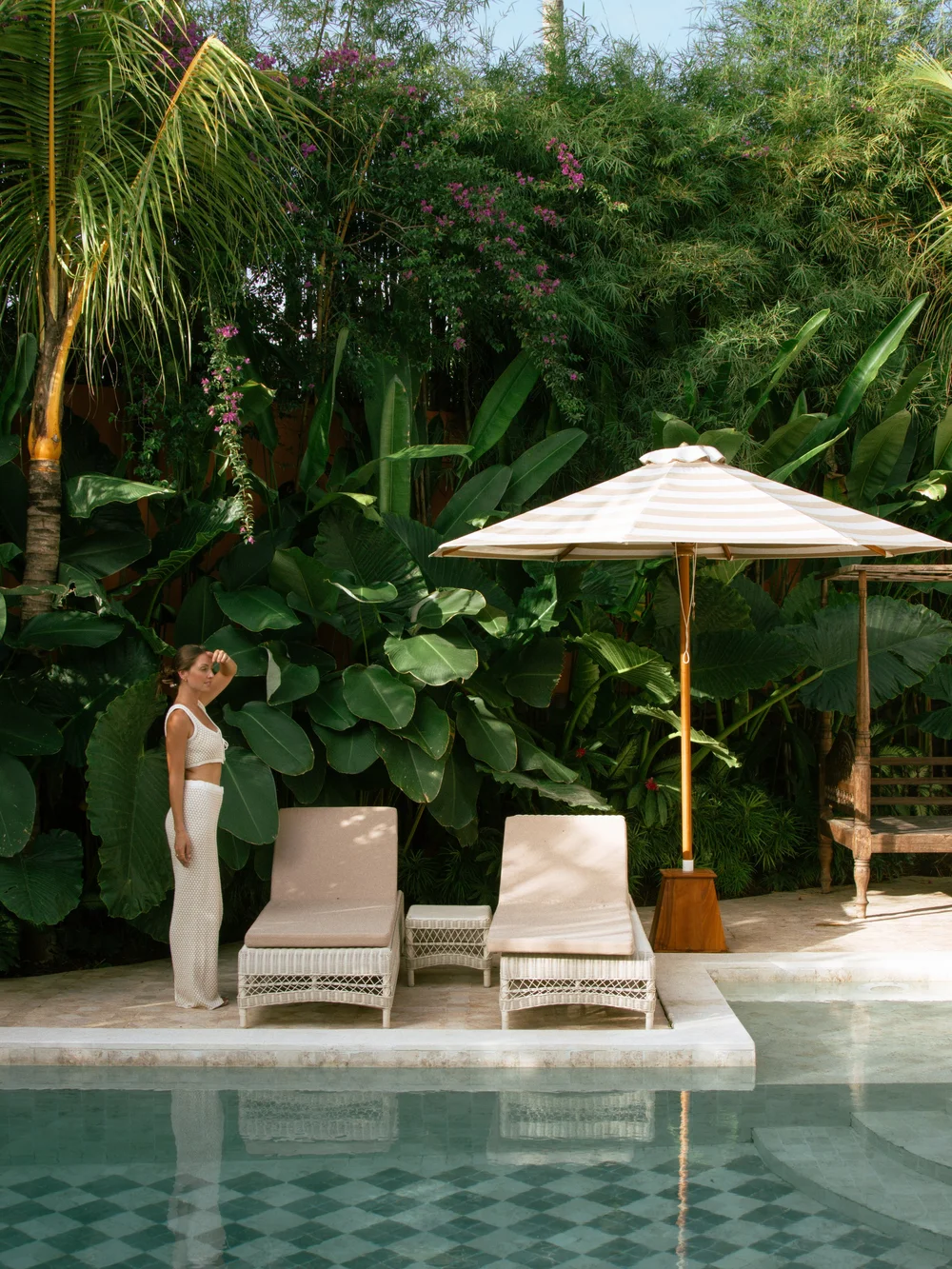 A woman stands beside two lounge chairs by a pool, surrounded by lush tropical plants and a parasol.