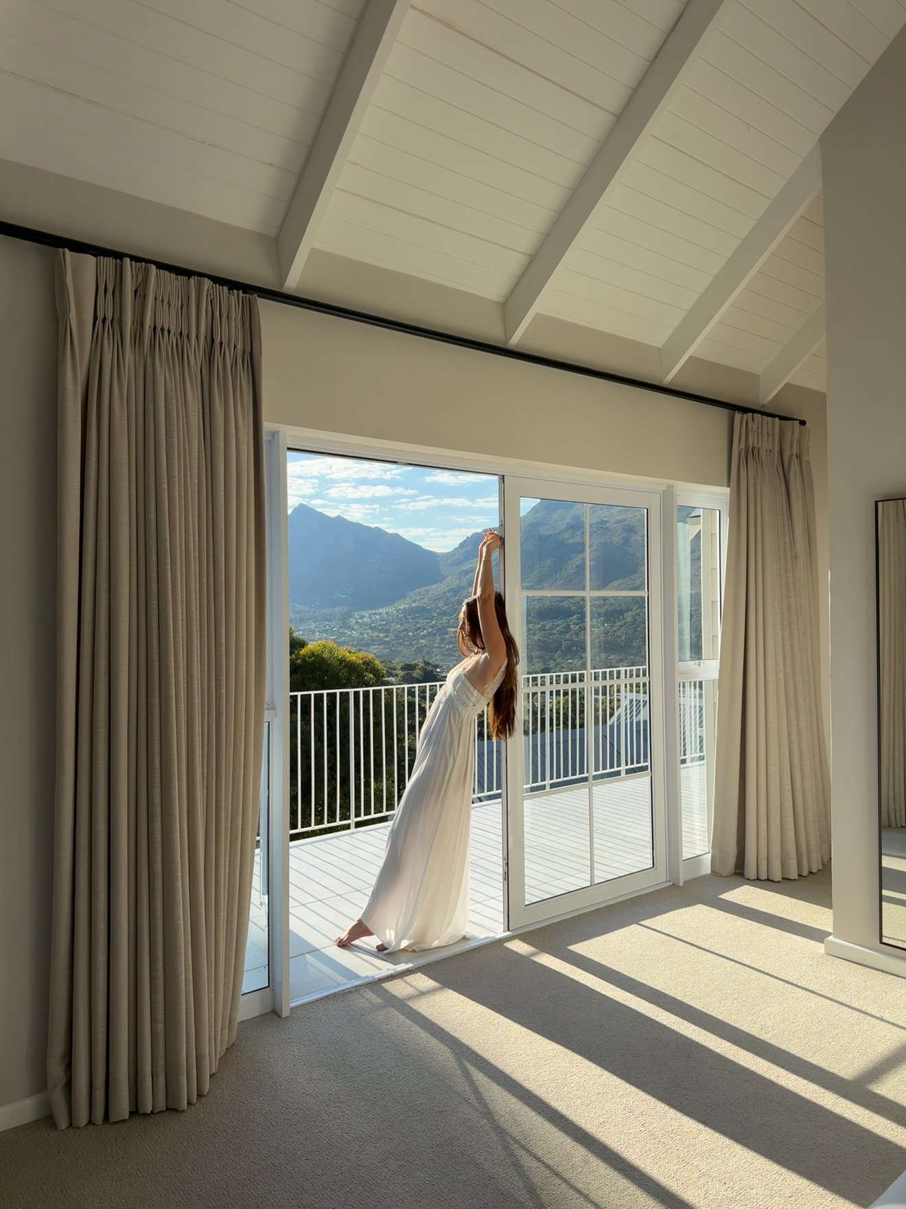 A woman in a flowing white dress stands by an open sliding door, overlooking a scenic mountain view.